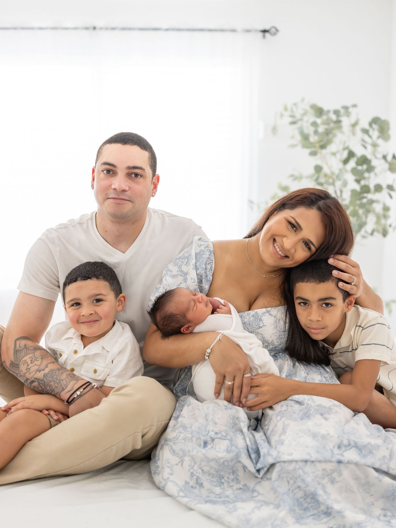 A family relaxes together on a white bed, with a newborn baby and light colored decor in the background.
