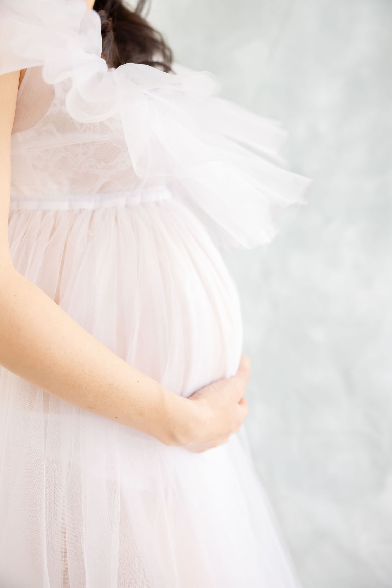 Side view of a pregnant belly draped in flowing white chiffon maternity dress against light background.