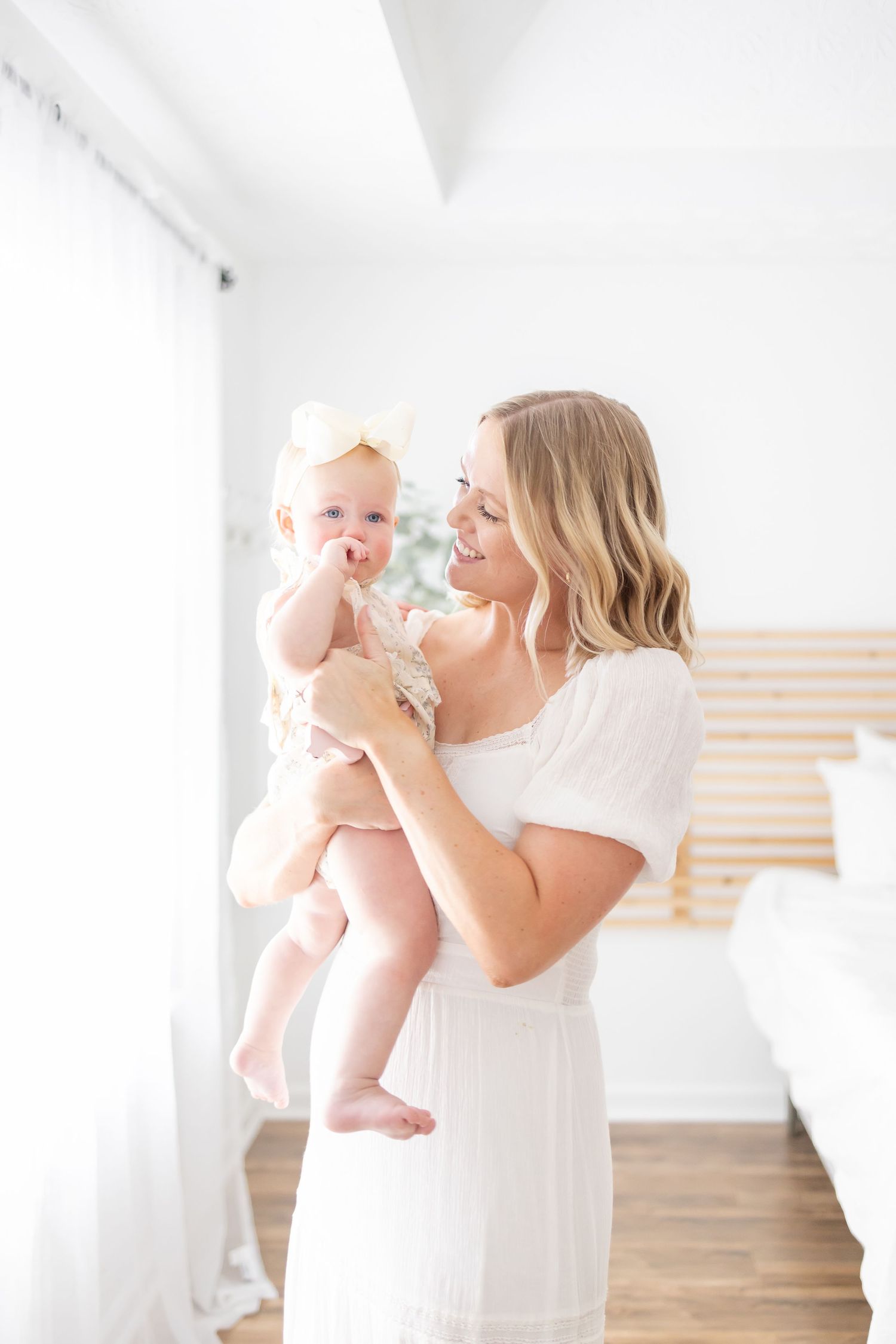 A parent in a white dress holds a small baby in the air while standing in a bright minimalist room with wood floors.