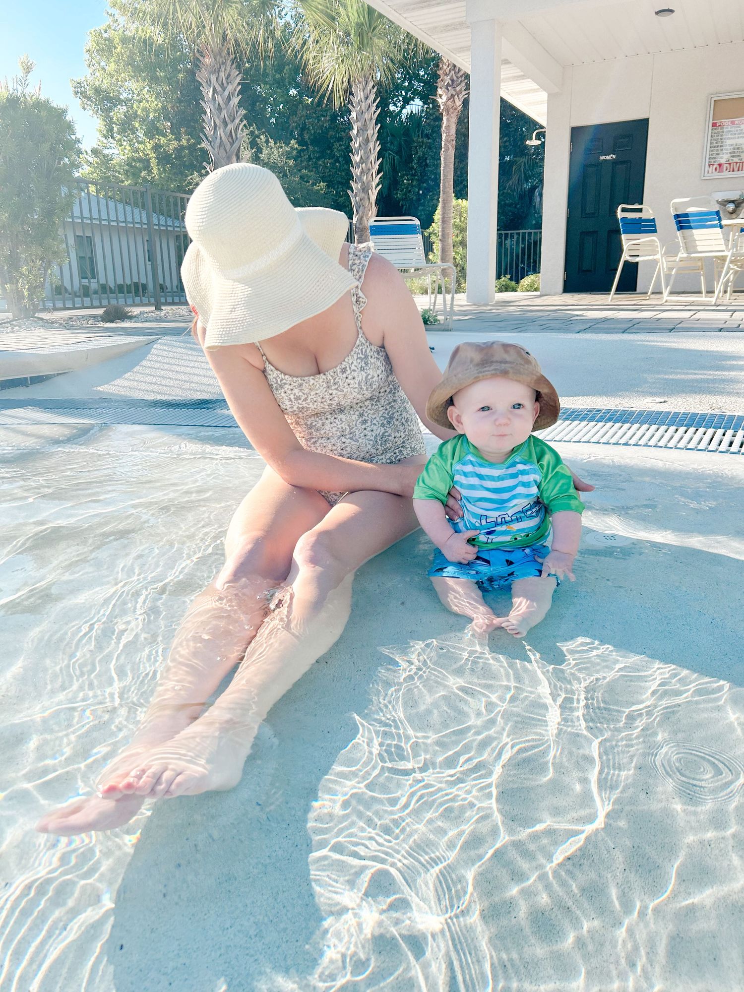 A toddler in blue and green swimwear splashes and plays in shallow pool water on a sunny summer day.