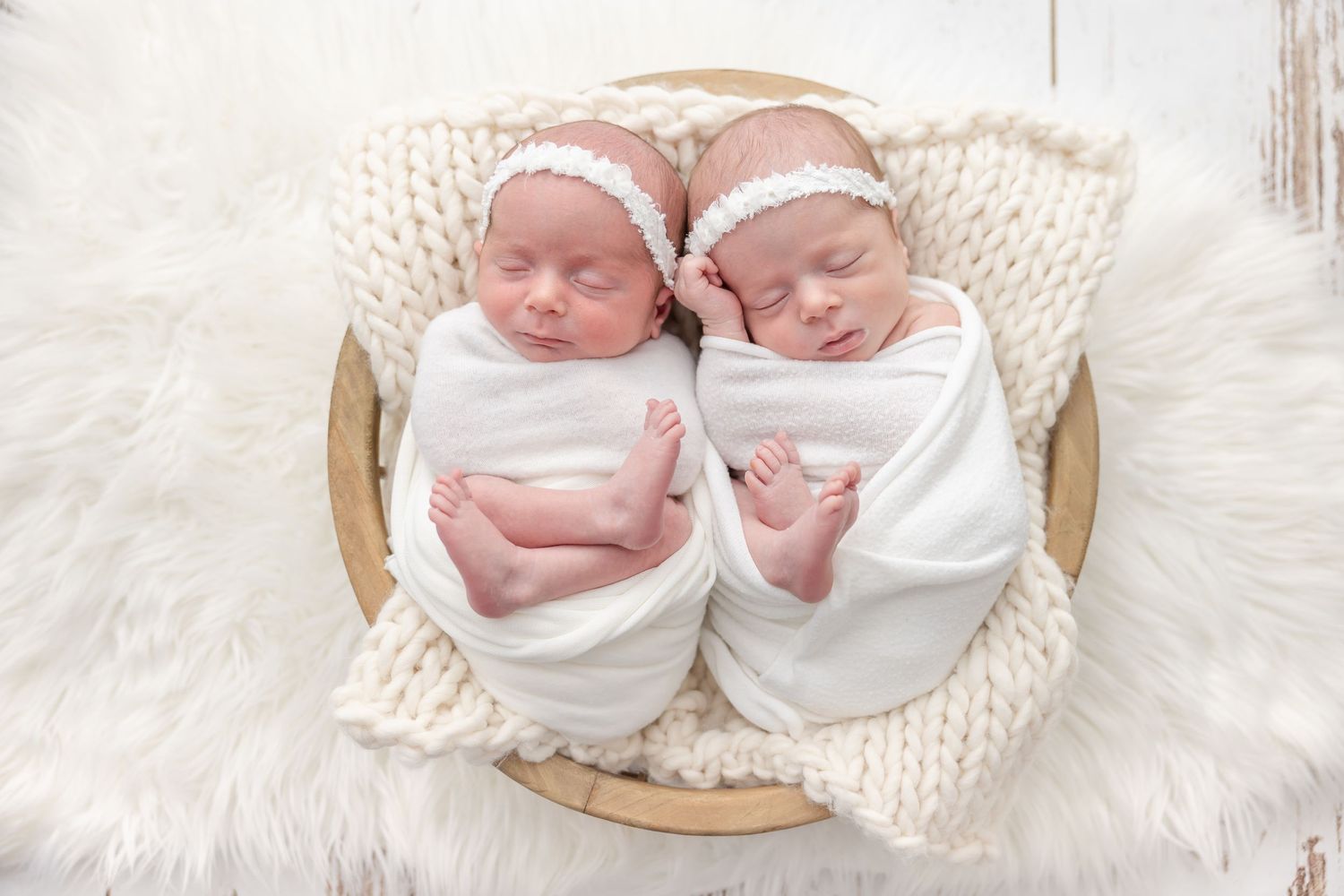 Two swaddled newborn babies wearing matching white headbands rest together on soft white textured blankets.