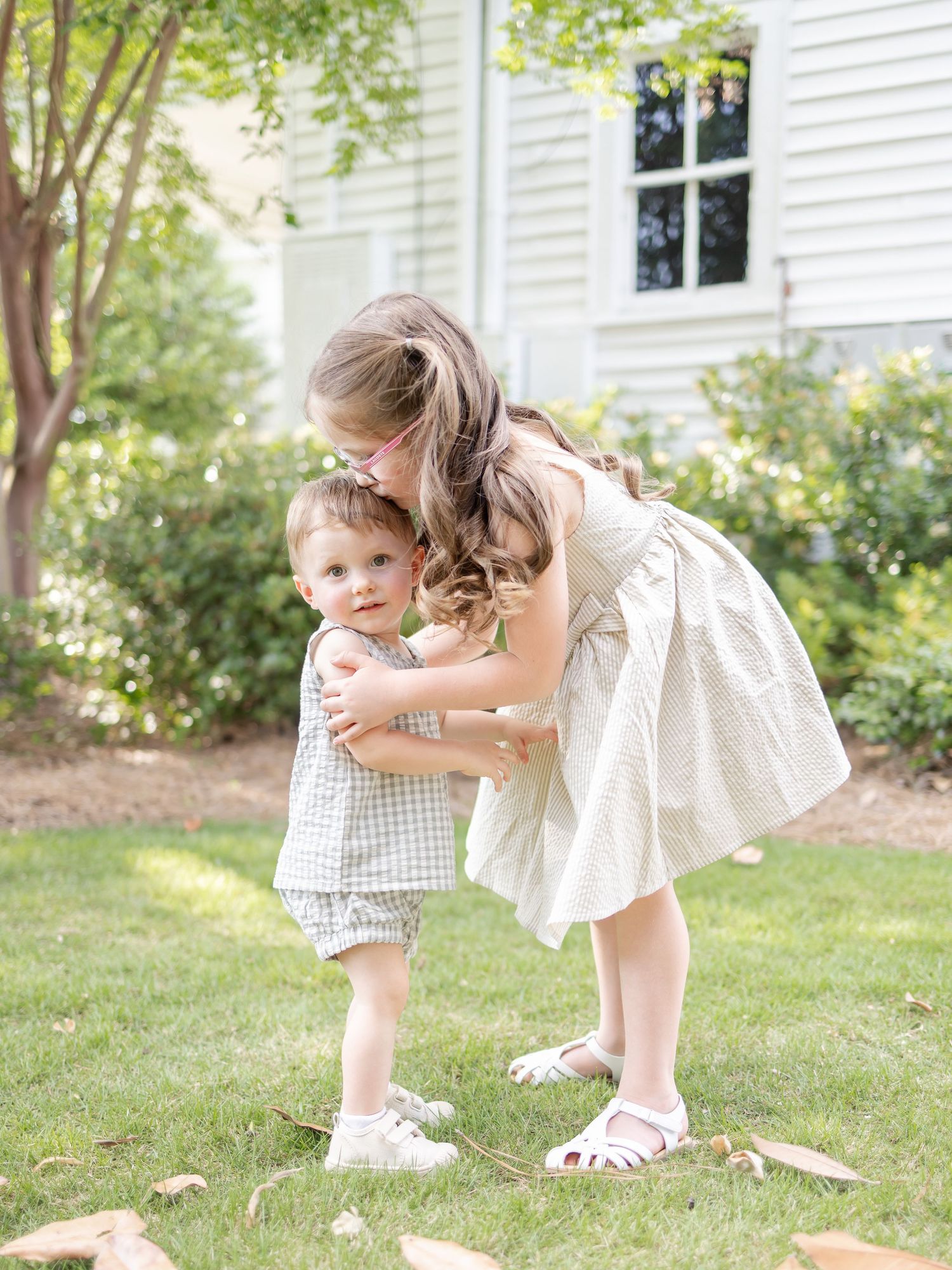 Children in coordinating light-colored outfits play together on a sunny lawn beside a white house with greenery.