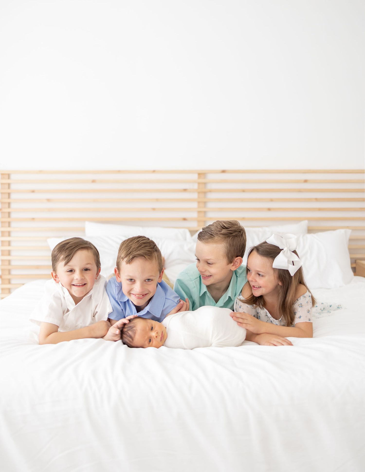 Young children laying on white bedding while admiring a baby in a peaceful bedroom setting.