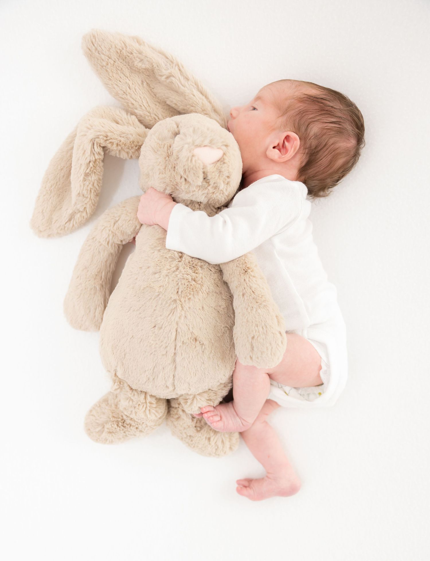 Baby in white sleeper cuddles with large plush beige bunny rabbit toy on white background.