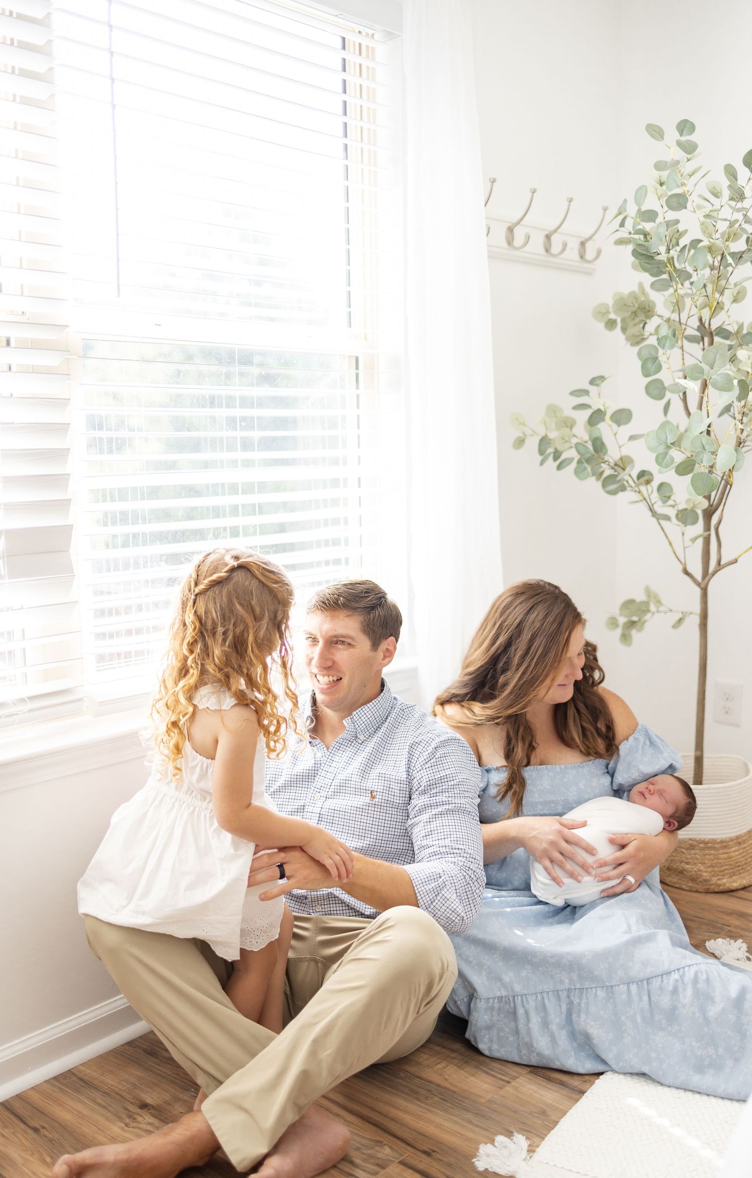 A bright modern family sits together on a hardwood floor by a window with a decorative plant in a cozy living room.