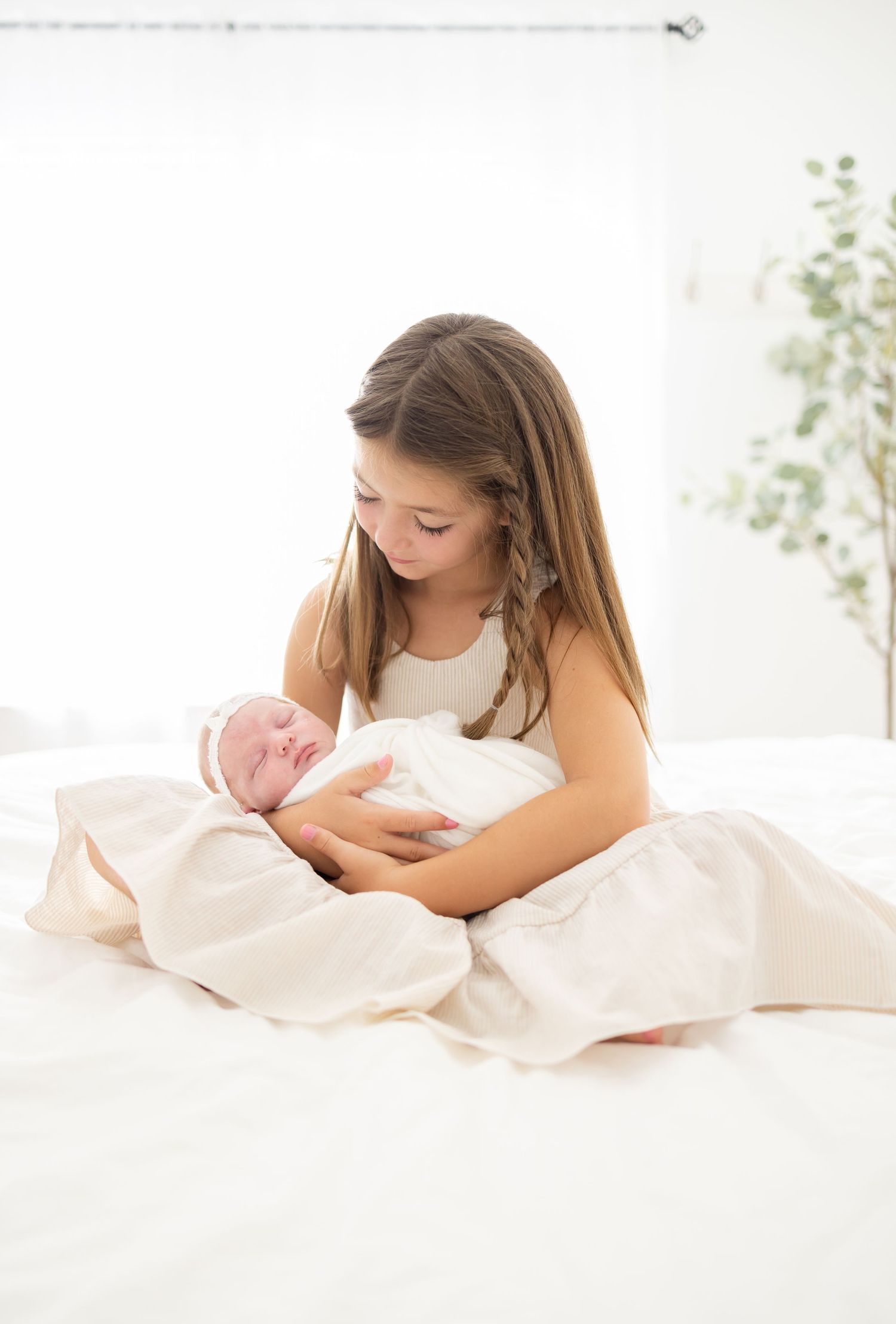 A young girl in white sits on a bed holding and gazing lovingly at a newborn baby wrapped in soft fabric.