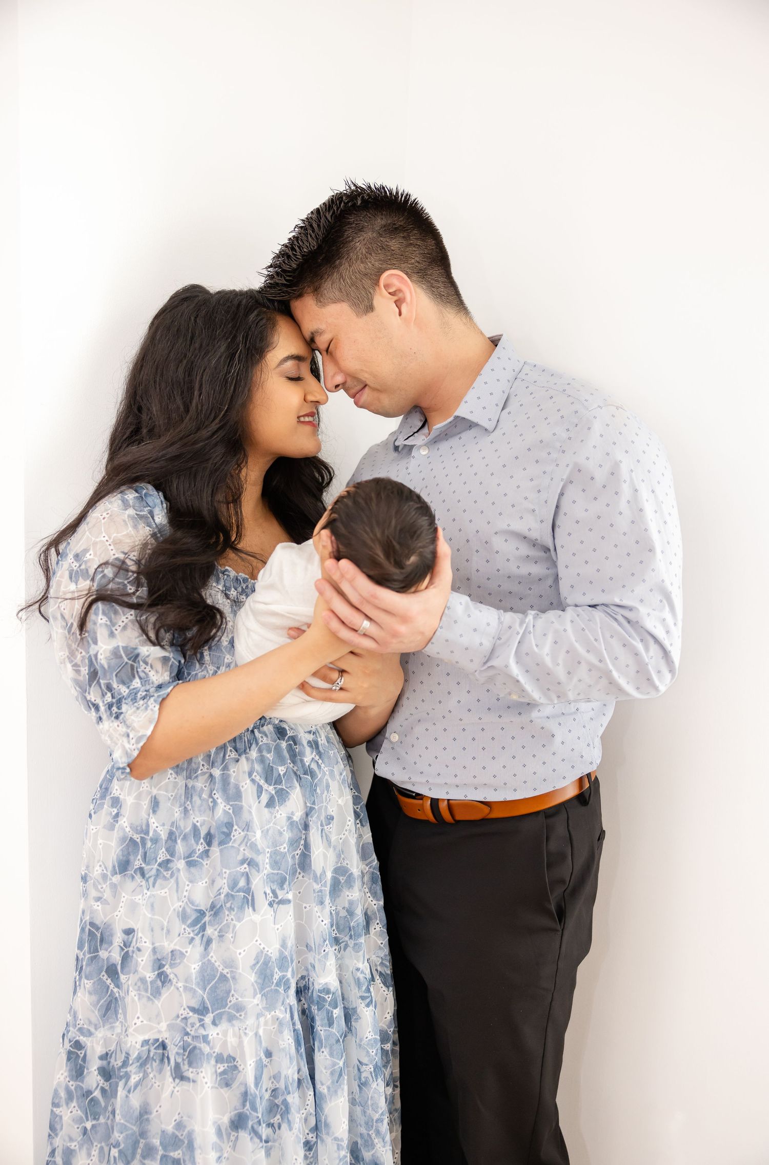A couple in coordinated blue and gray outfits share a tender moment while holding their newborn baby against a white backdrop.