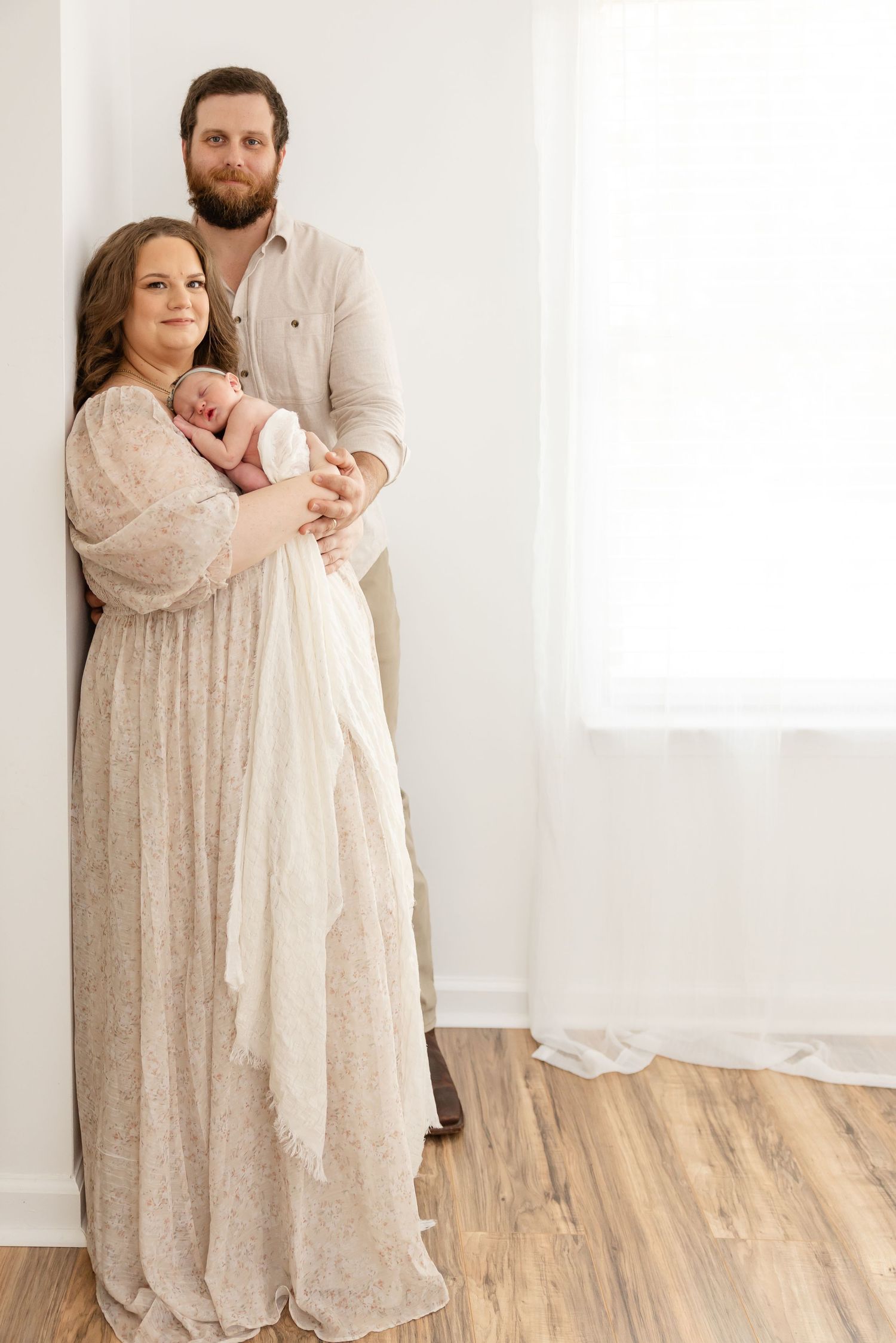 A family stands together near a bright window in their home wearing soft neutral-colored clothing.