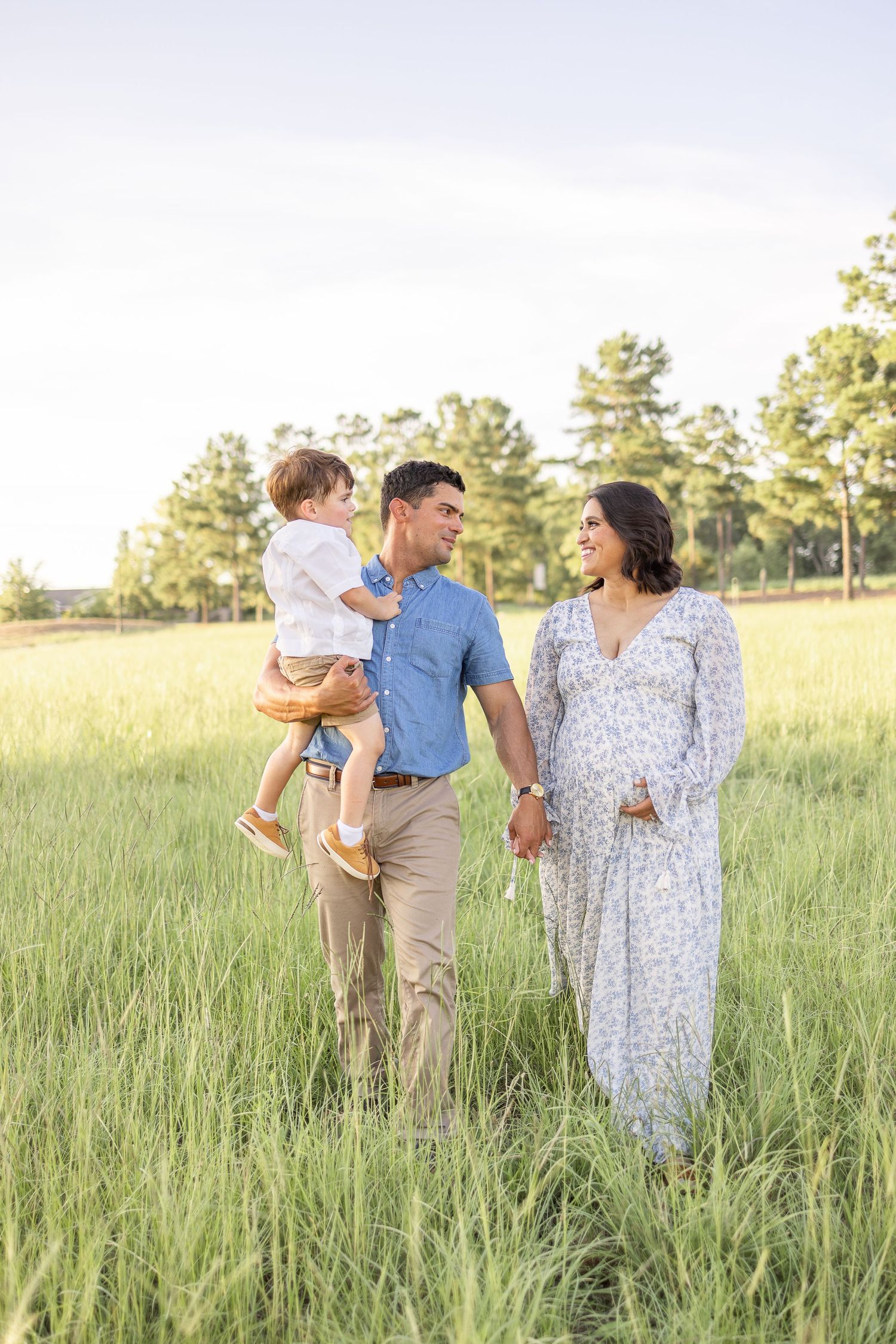 A family stands together in a sunny meadow with tall grass and pine trees in the distance.