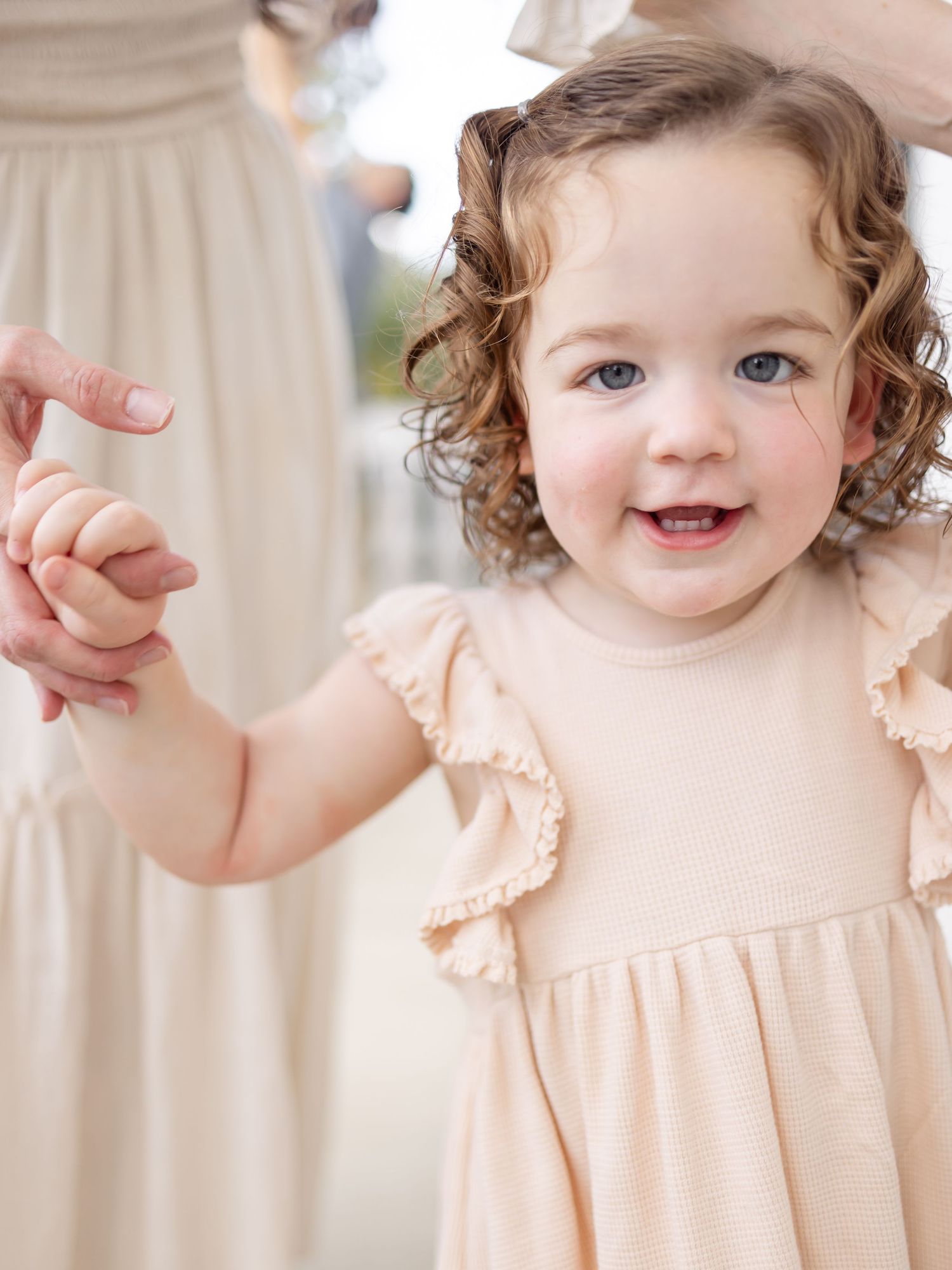 A young child in a peach-colored ruffled dress smiles brightly while holding someone's hand.