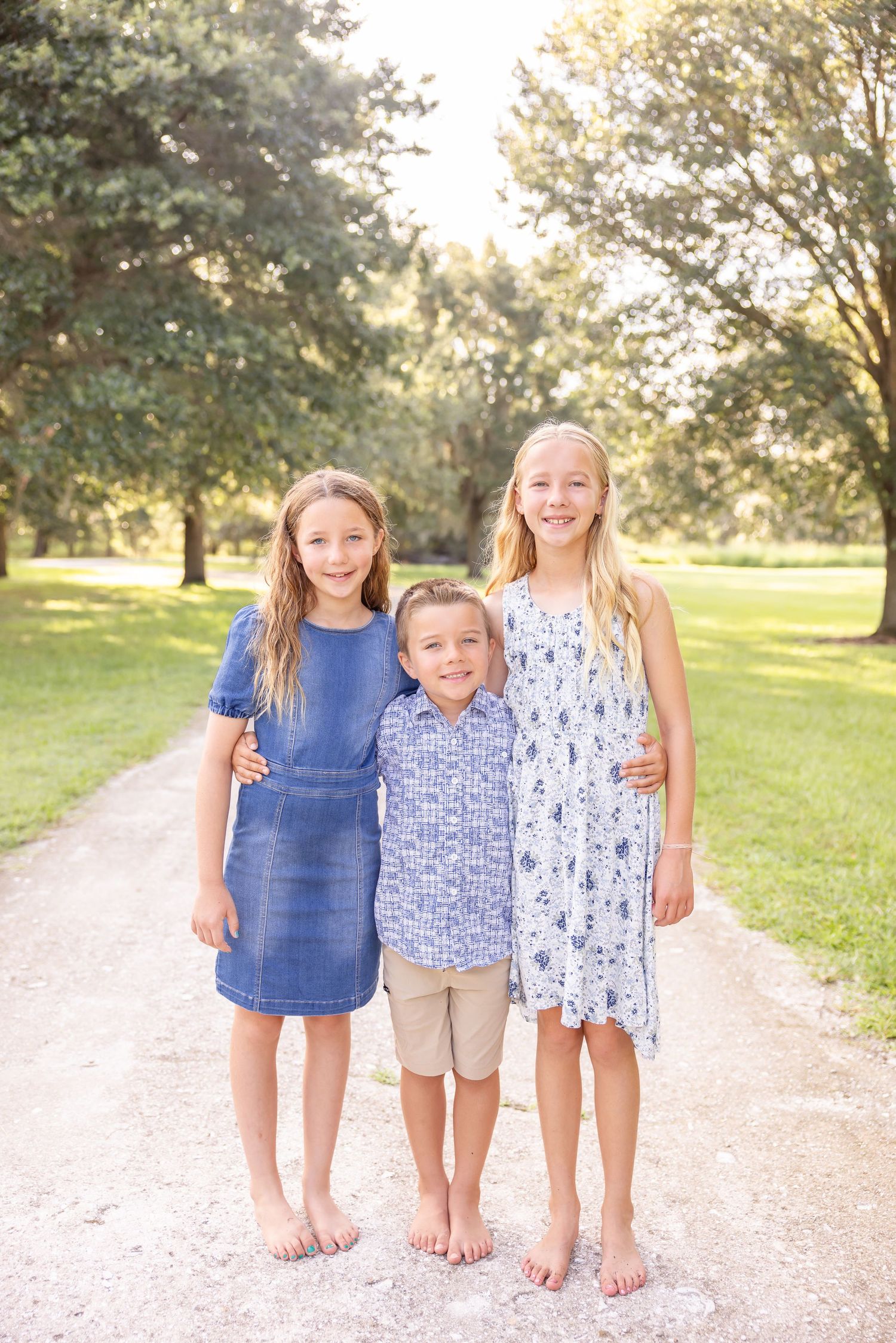 Three children in summer attire stand barefoot on a gravel path surrounded by sunlit trees and grass in a park.