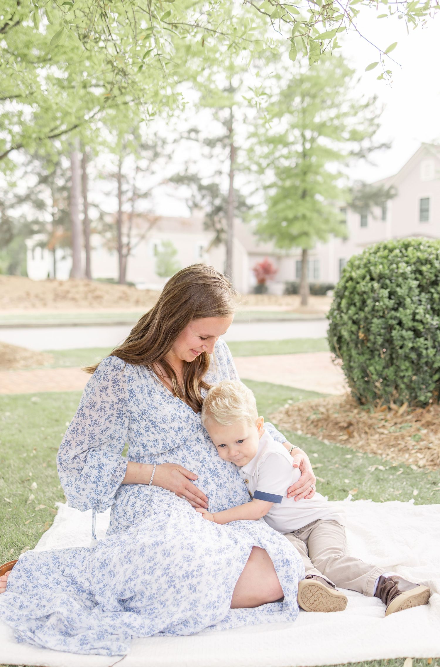 Parent and small child sit together reading on white blanket in sunny garden setting with bushes and trees.