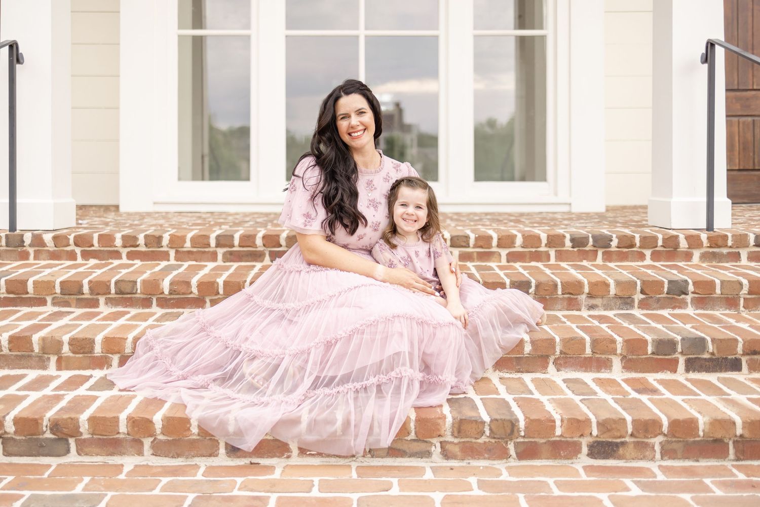 A mother and daughter wearing matching pink tulle dresses sit together on brick steps in front of a white building facade.