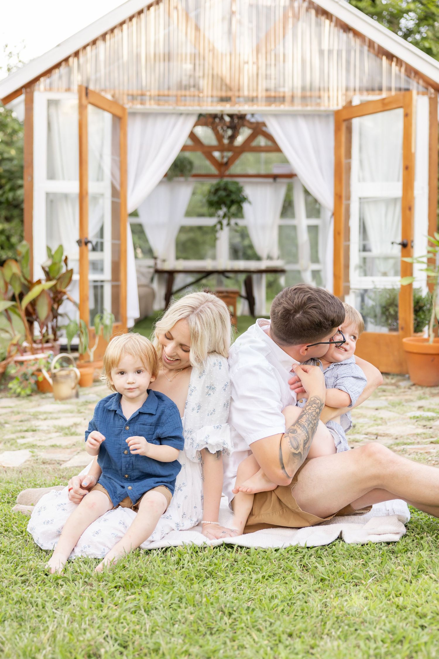 Family relaxes on blanket in front of rustic greenhouse with white curtains and wooden beams in outdoor garden setting.