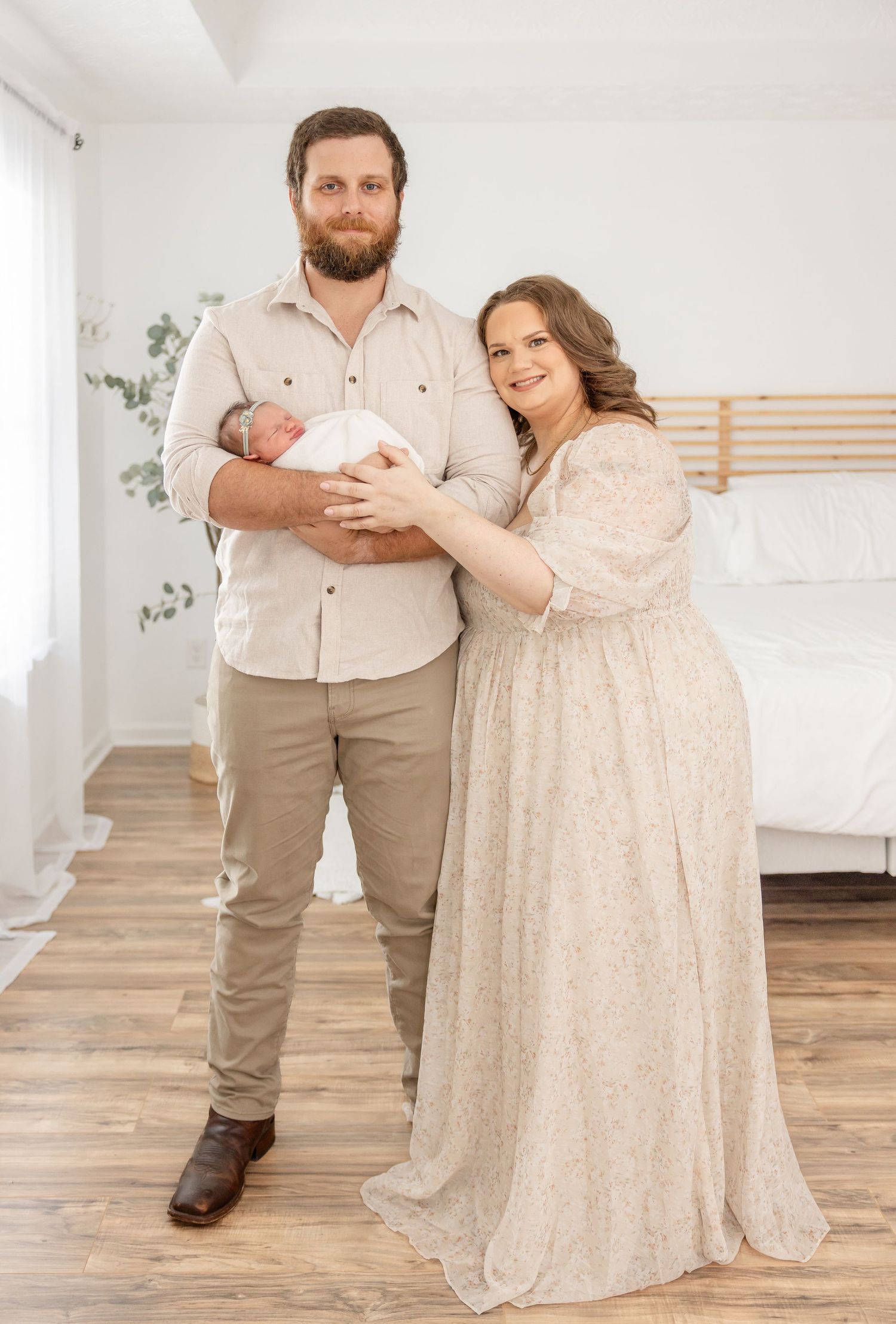 A couple in neutral tones stands in a bright bedroom holding their newborn baby, with wooden floors and minimalist decor.