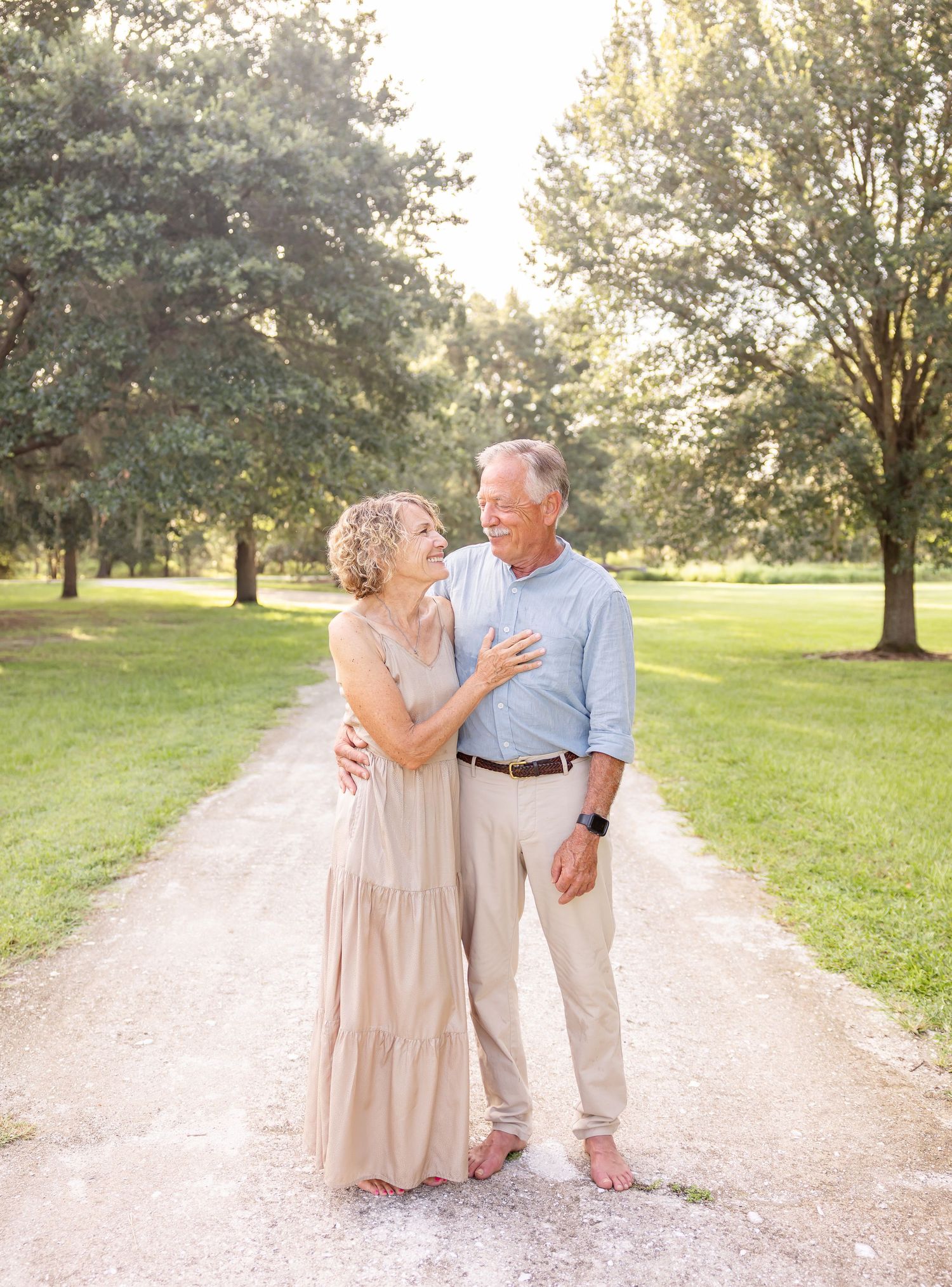 Couple embraces while standing on a sunlit gravel path surrounded by green trees in a serene park setting.