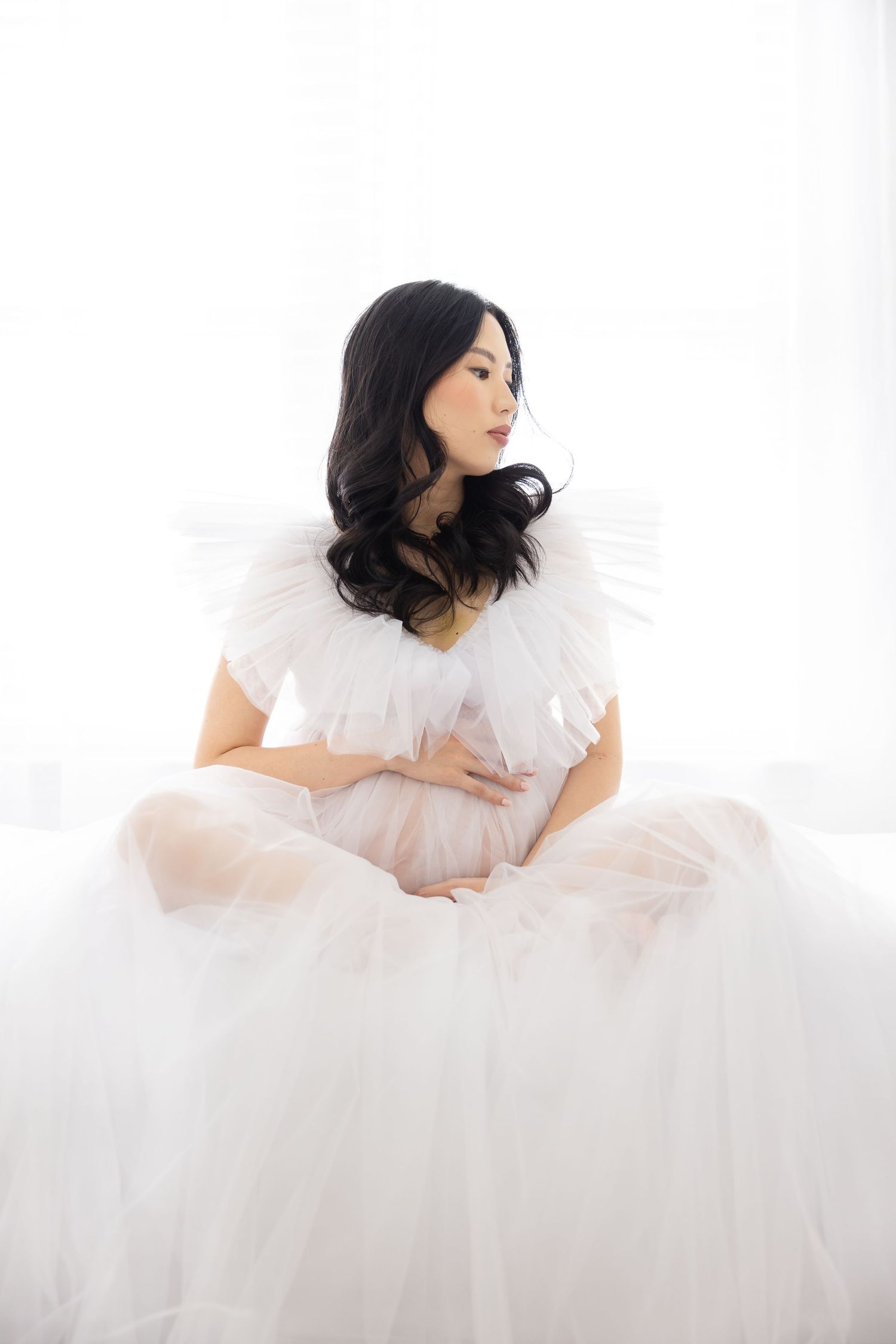 A person in an ethereal white tulle gown sits gracefully against a bright background, creating a dreamy bridal portrait.