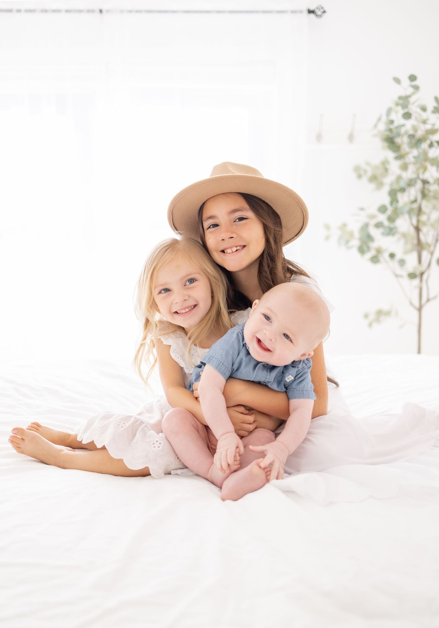 Three children sitting together on white bedding, with one wearing a tan hat and another in blue clothing.