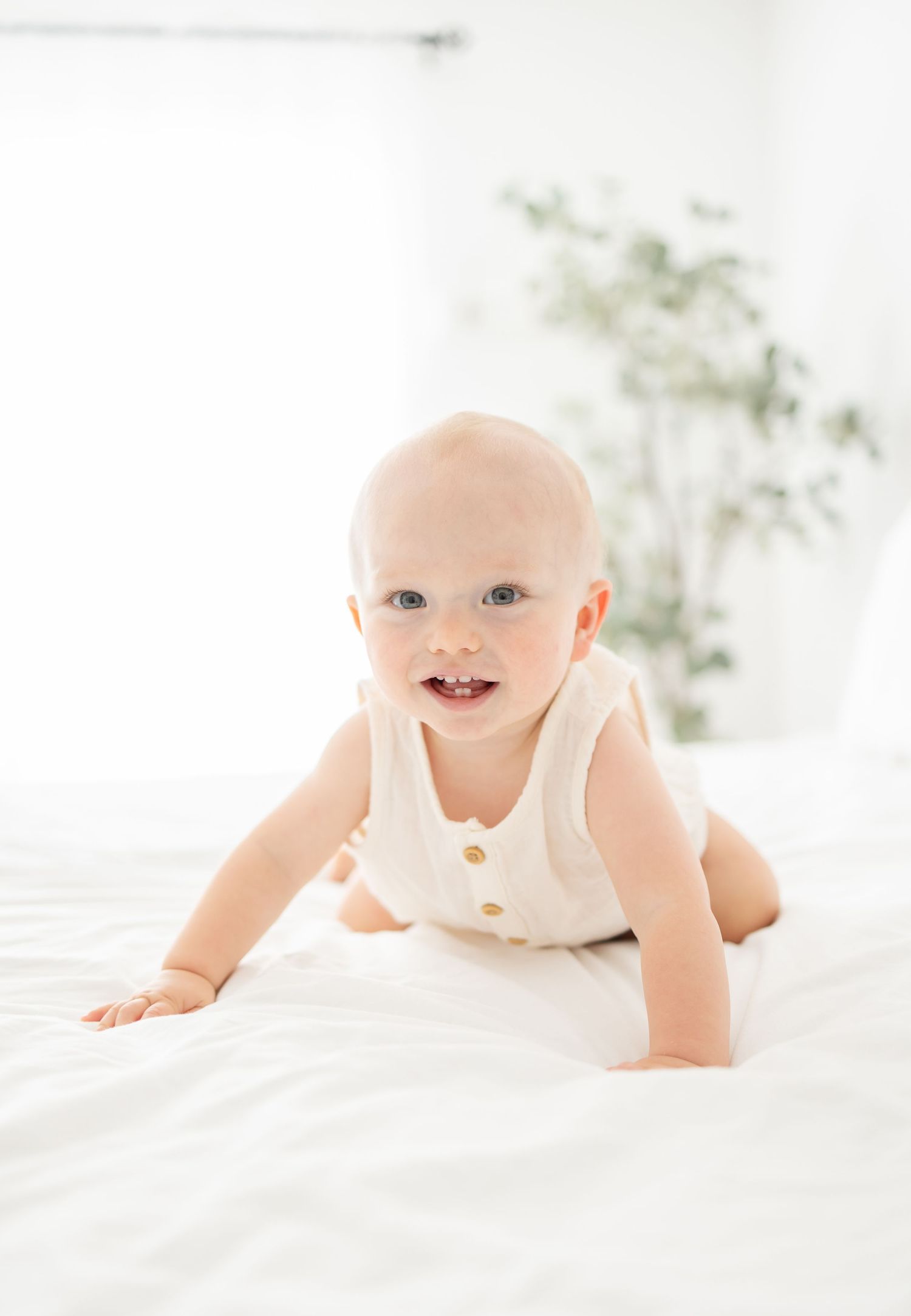 Sweet baby in white onesie crawls and smiles on soft white bedding in bright, airy setting.