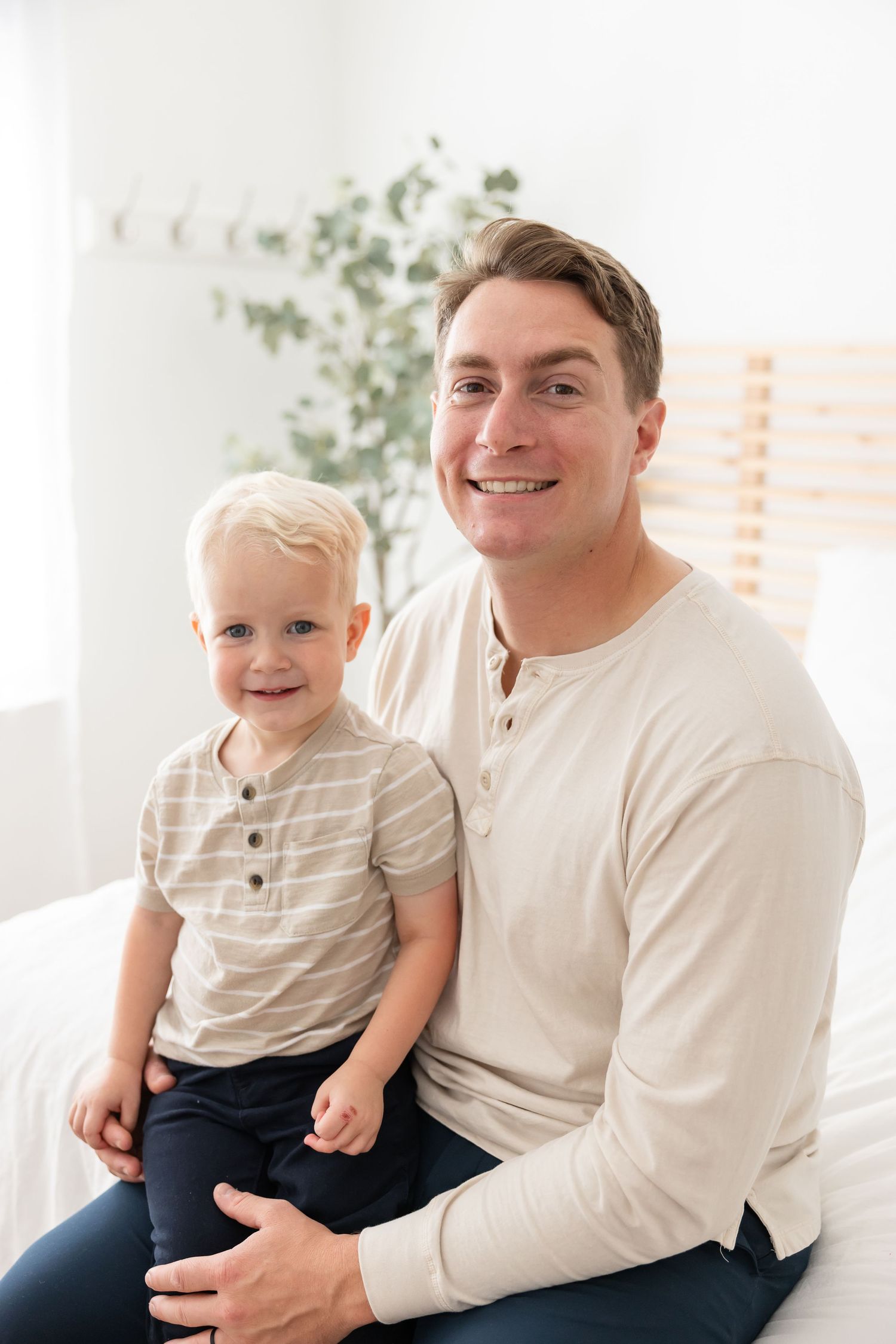 A parent and young child sit together indoors, both wearing light colored shirts and sharing smiles.