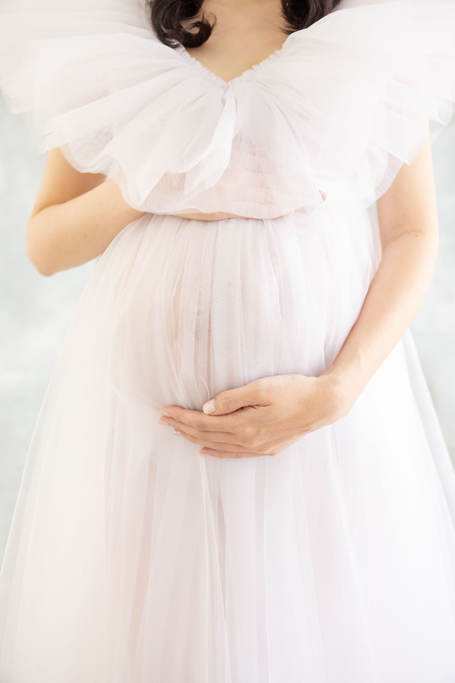 Ethereal maternity photoshoot featuring a flowing white dress against soft neutral background, emphasizing the baby bump.