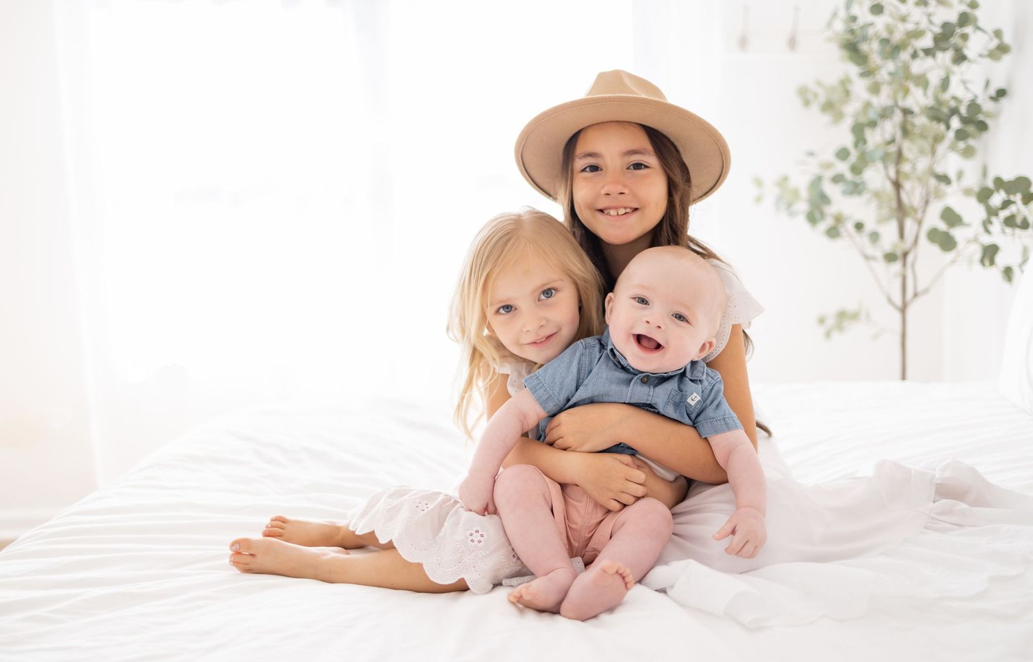 Three children sit together on white bedding while wearing summer clothing and a straw hat.