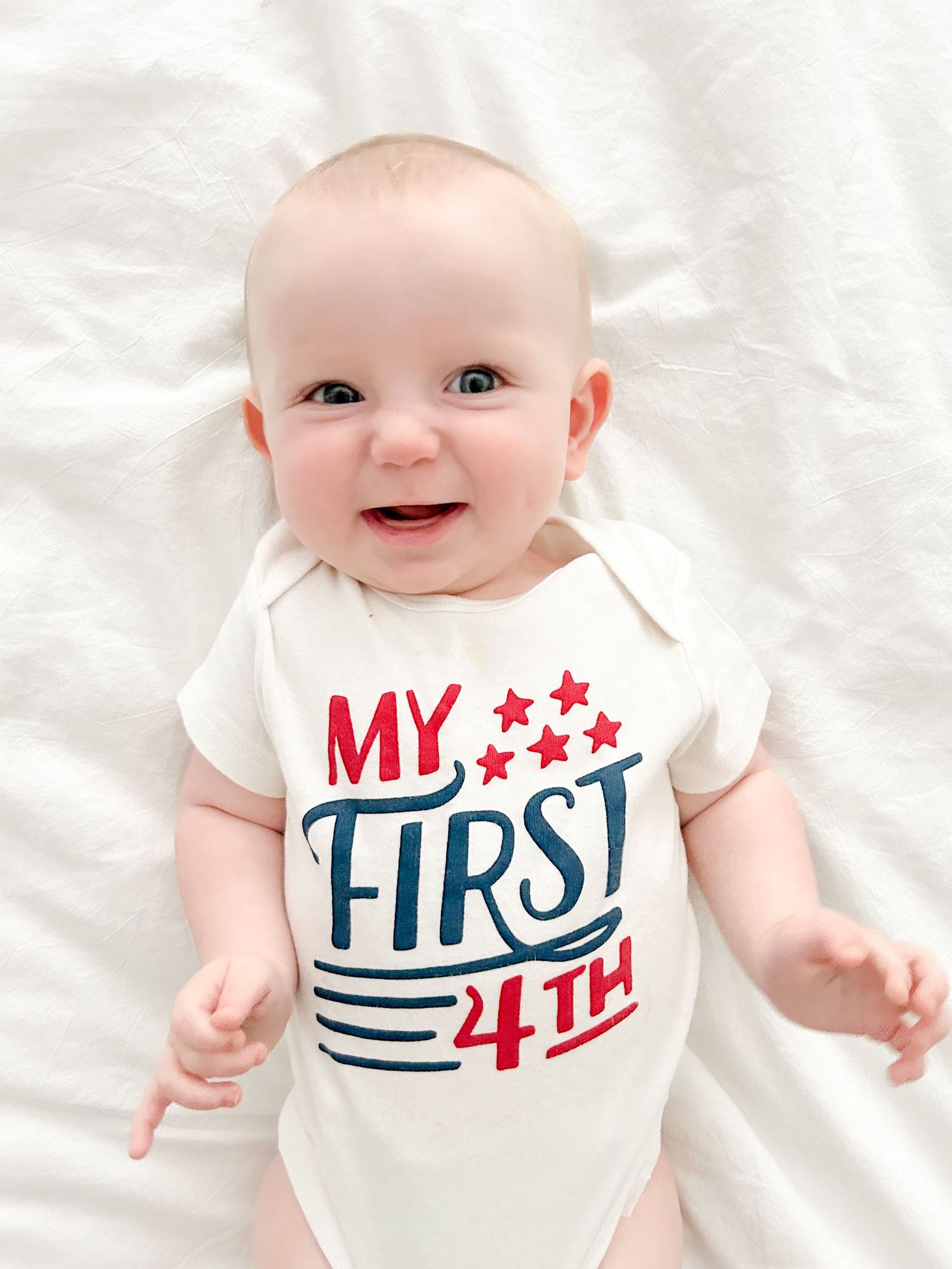 Baby wearing white onesie with patriotic 'My First 4th' text and stars design lying on white fabric.