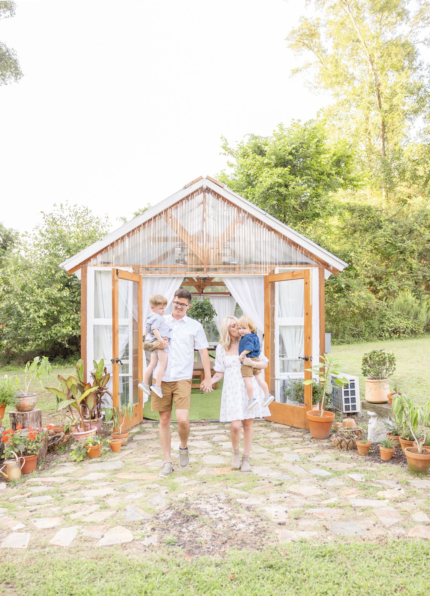 A rustic wooden greenhouse with potted plants and a beautiful stone pathway surrounded by lush greenery on a sunny day.