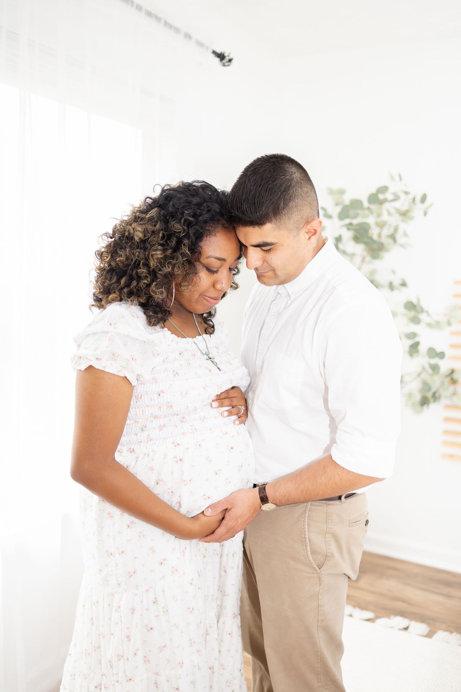 An expecting couple in white and cream outfits share an intimate moment while cradling her baby bump in a bright interior.