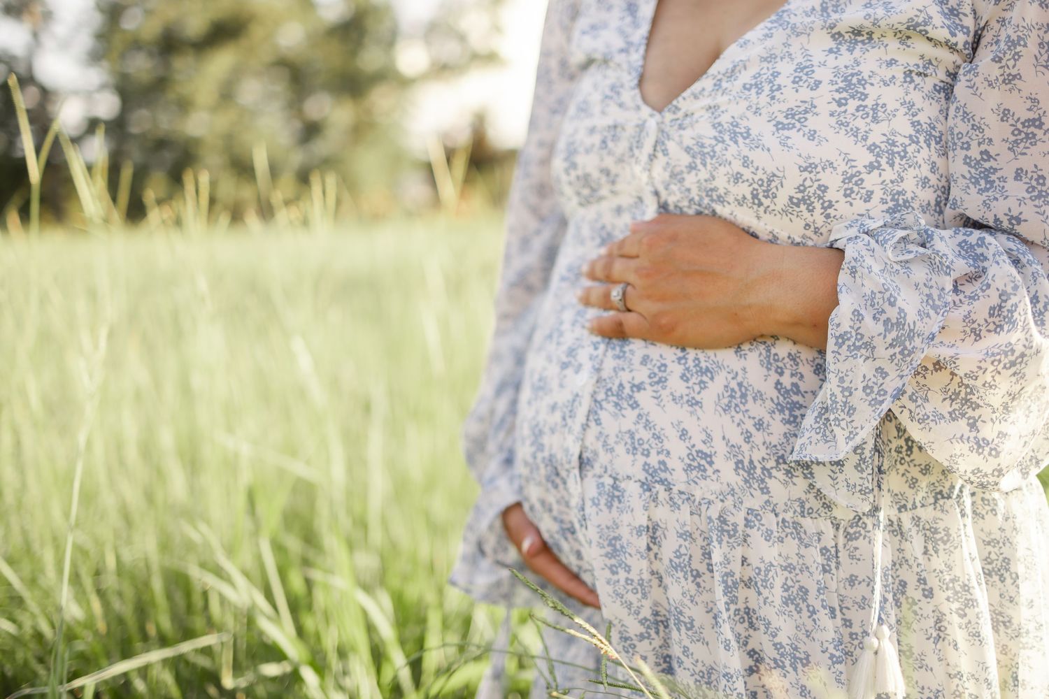 A pregnant person wearing a floral dress stands in tall grass during golden hour, gently cradling their belly.
