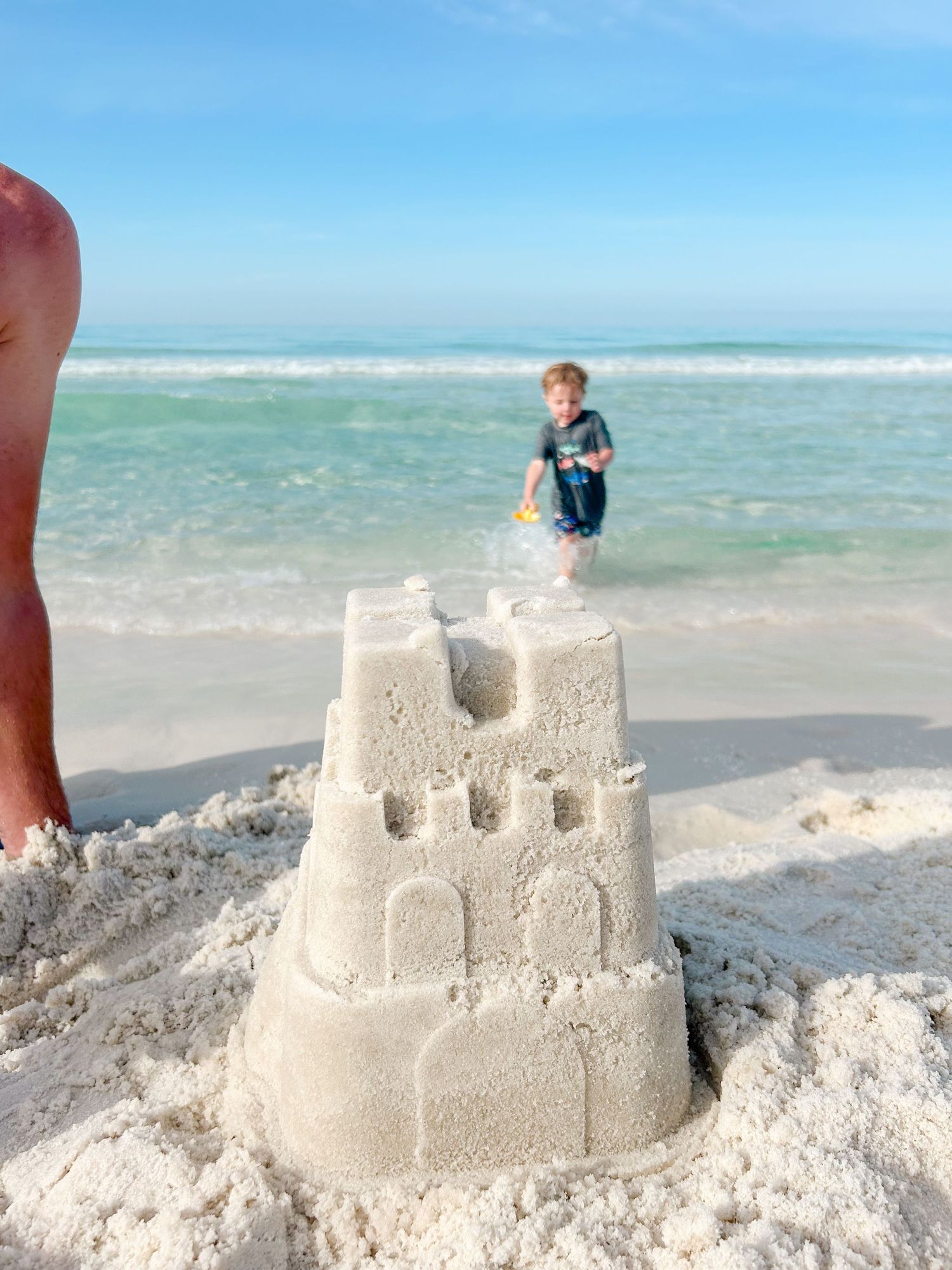 A detailed sandcastle on a white sandy beach with turquoise ocean waters and a person playing in the waves.