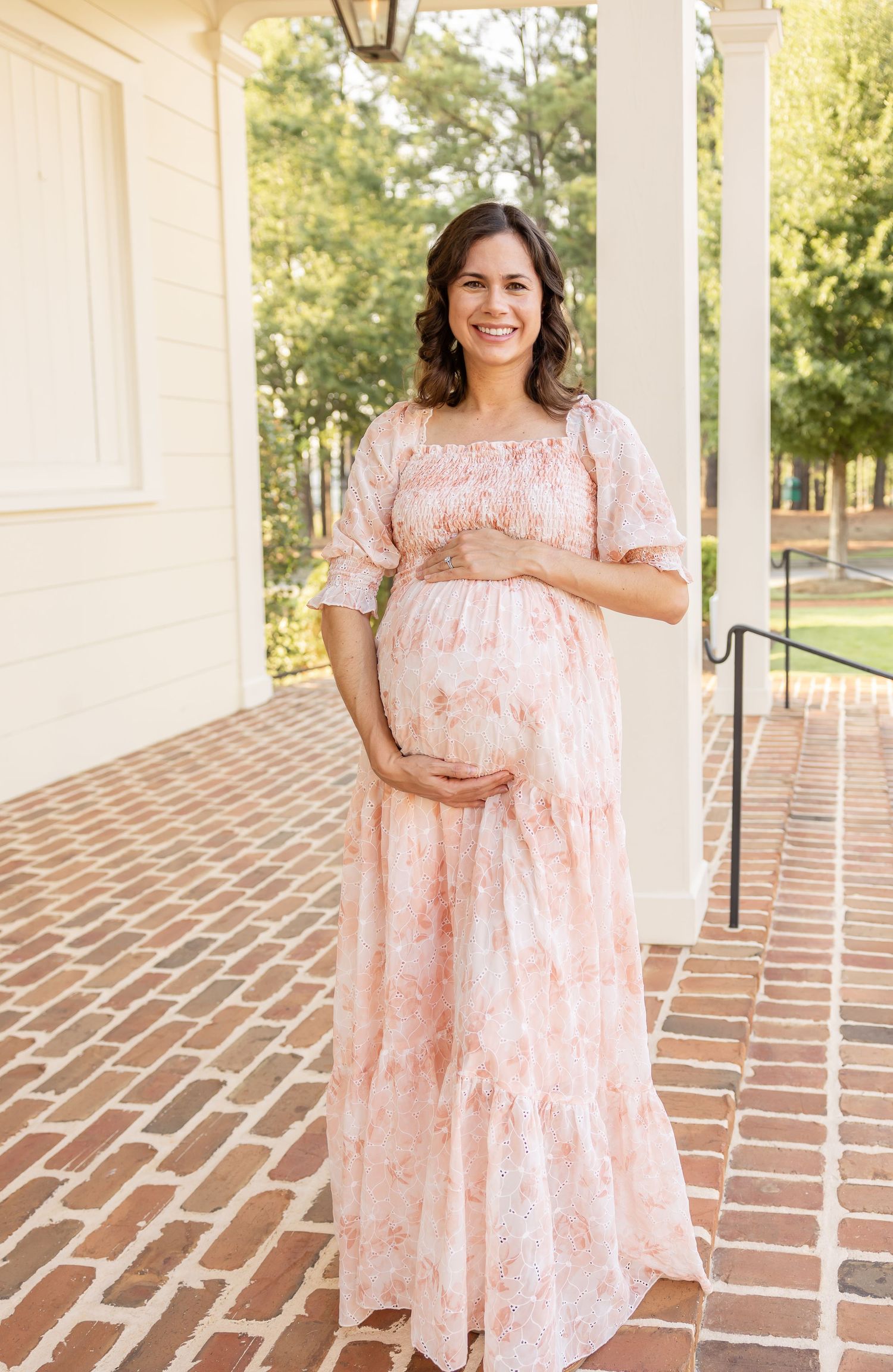 A person in a pink floral maternity dress stands on a brick porch with white columns on a sunny day.