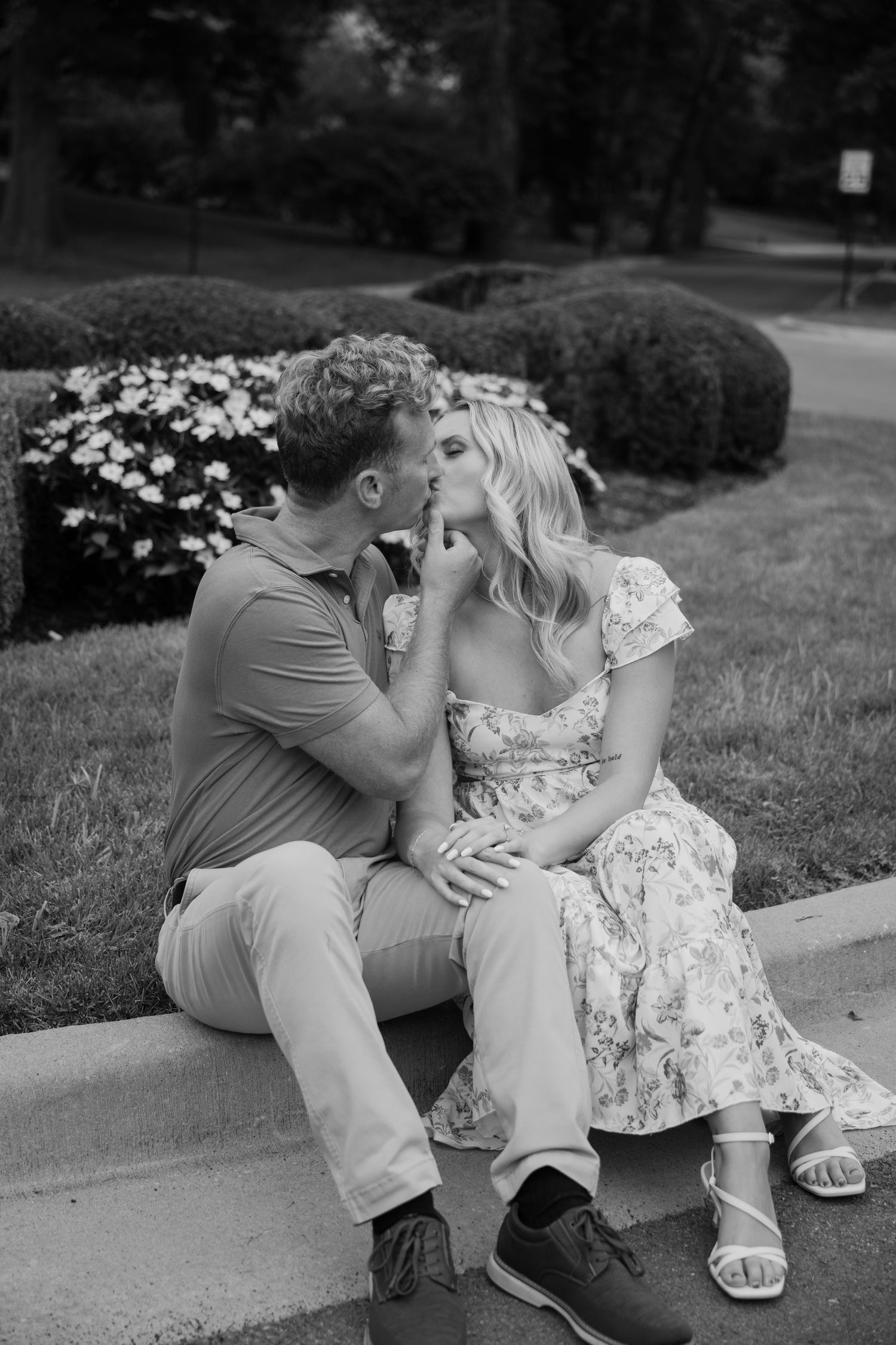 A romantic black and white photograph of a couple sharing a tender moment while sitting outdoors near blooming flowers.