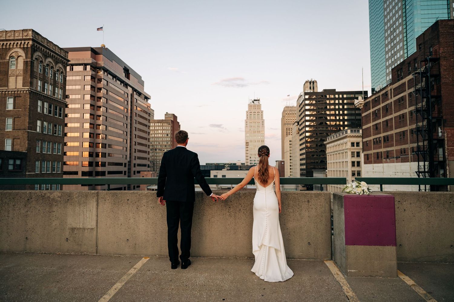 Couple in wedding attire standing on city rooftop at sunset overlooking urban skyline.