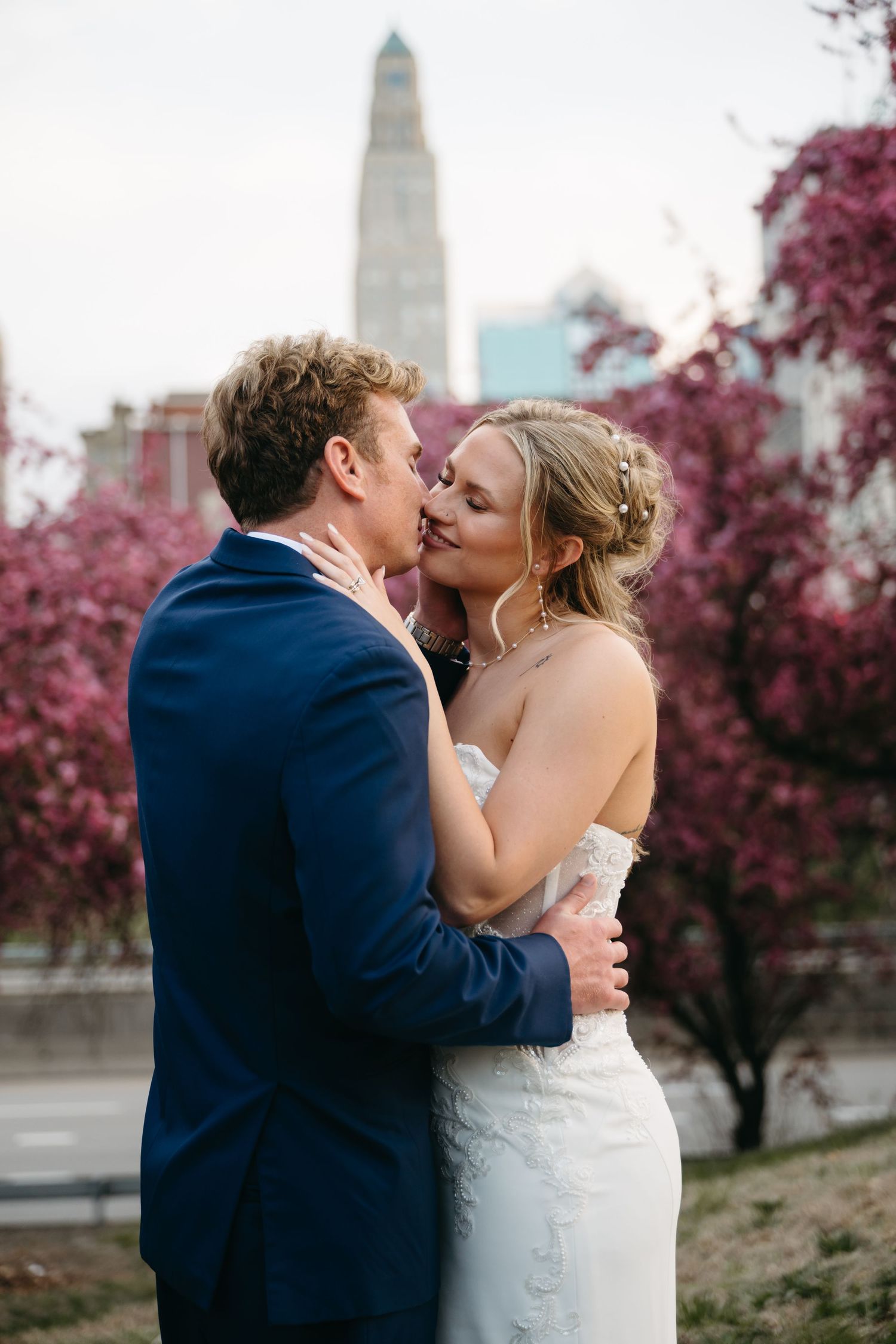 A romantic wedding couple shares a kiss with pink cherry blossoms blooming in the background.