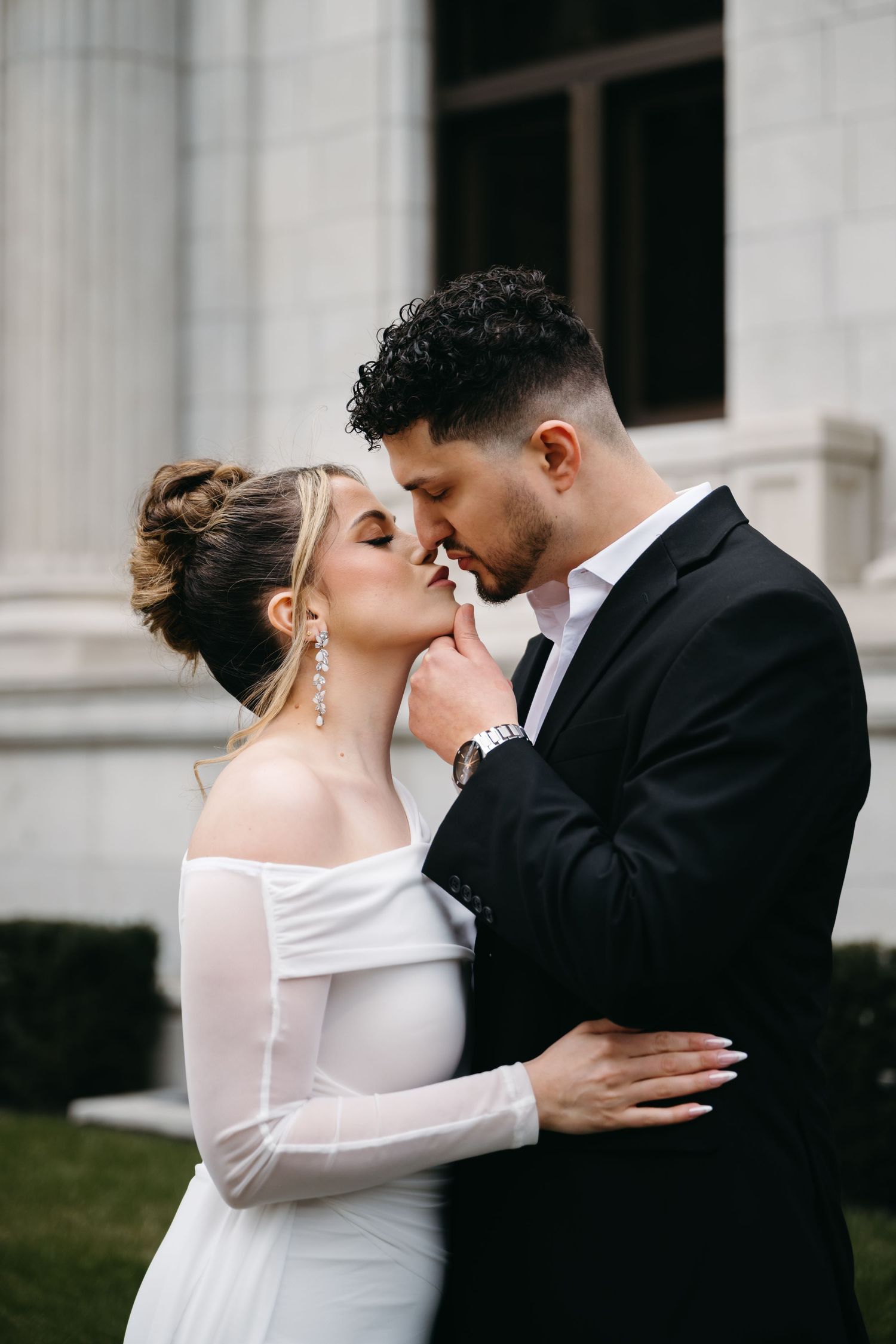 A romantic wedding moment with the bride in white long sleeve dress and groom in black suit embracing outside a building.