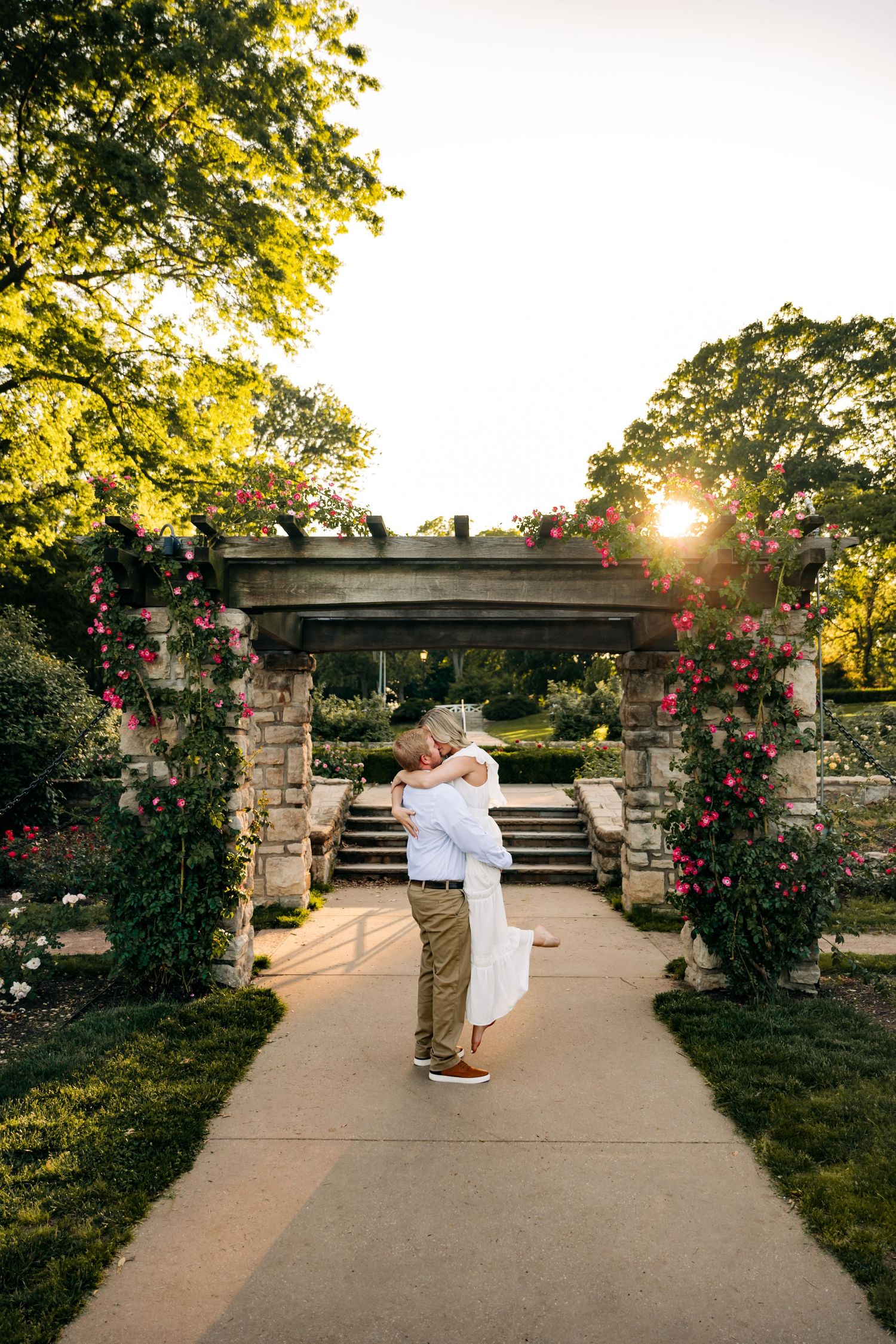 Couple embracing under a floral archway at sunset in a garden.