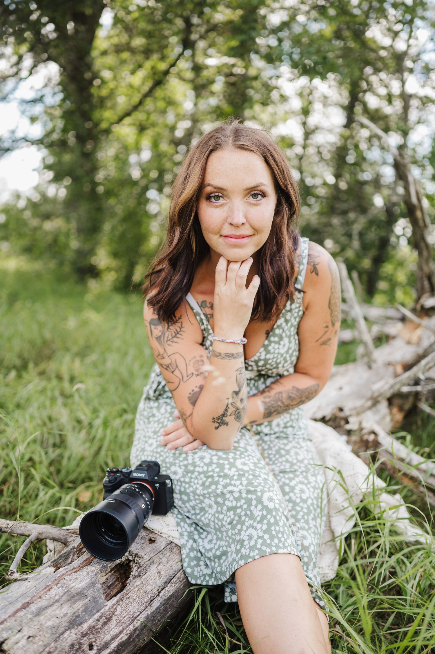 A portrait of a photographer with tattoos sitting on a log holding camera equipment in nature.