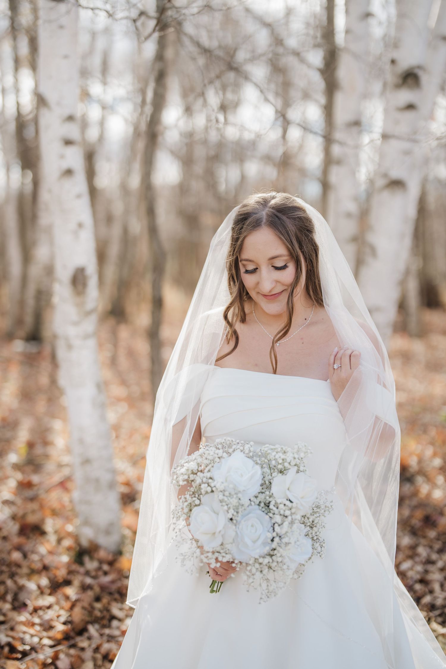A bride in a strapless white gown holds a delicate white bouquet while standing among autumn birch trees.