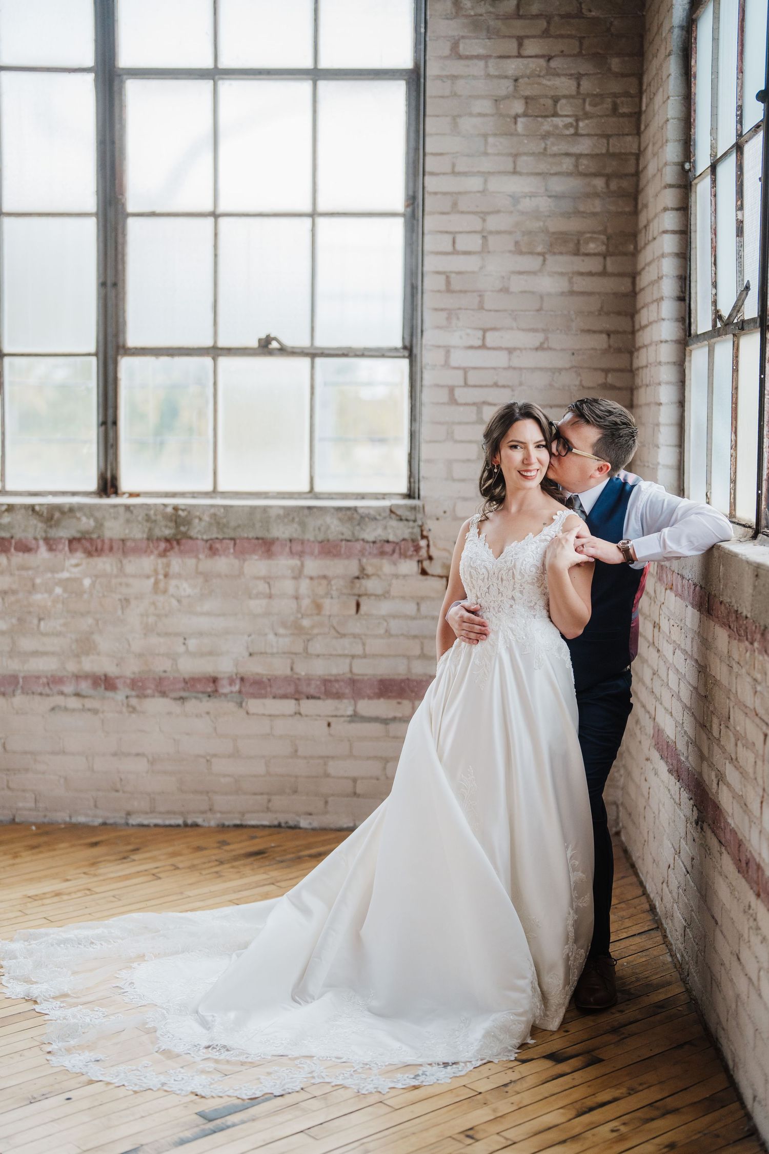 A romantic wedding couple embraces by large industrial windows in a brick loft setting.