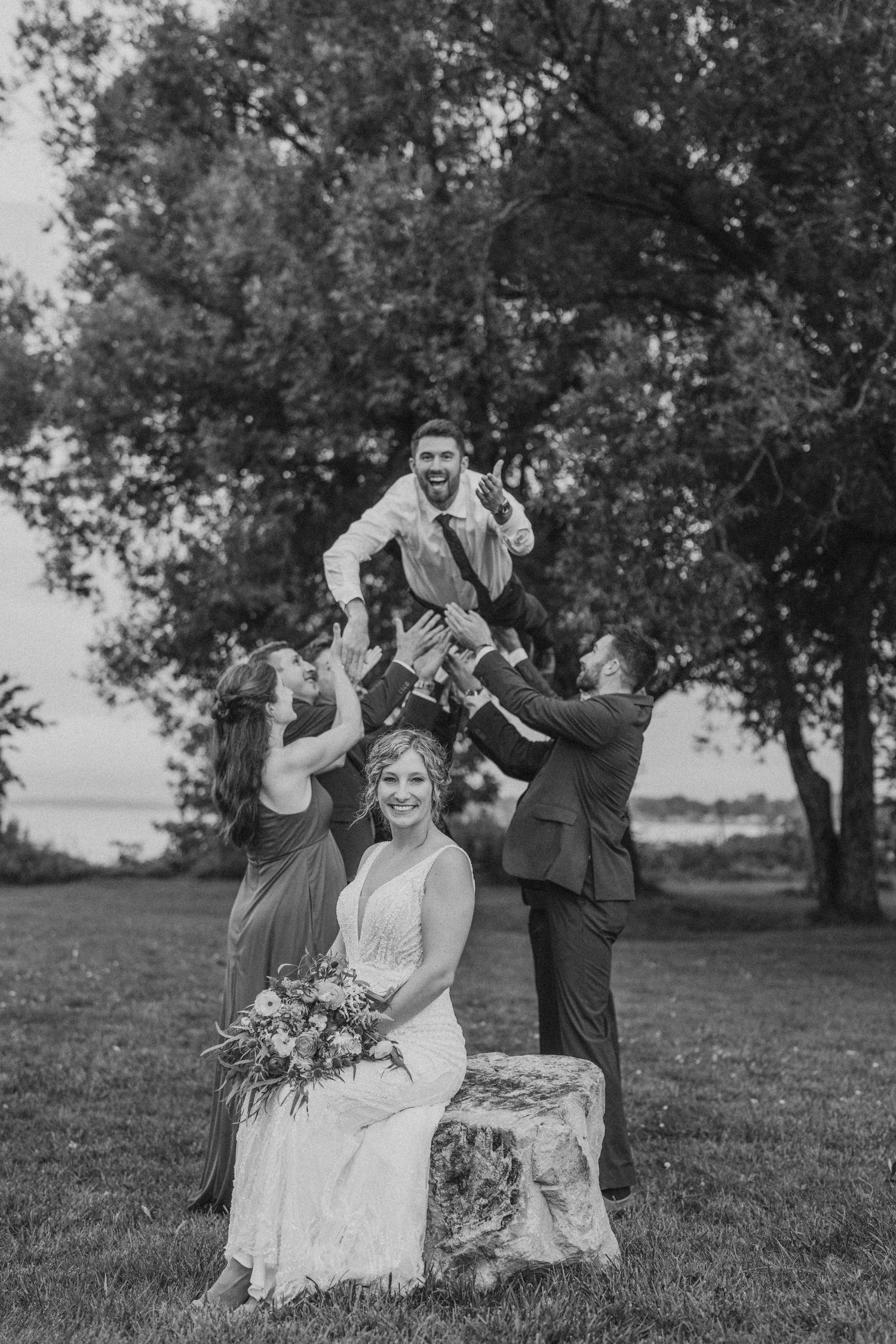 A fun wedding party photo with groomsmen lifting another while bride sits on vintage chair outdoors.