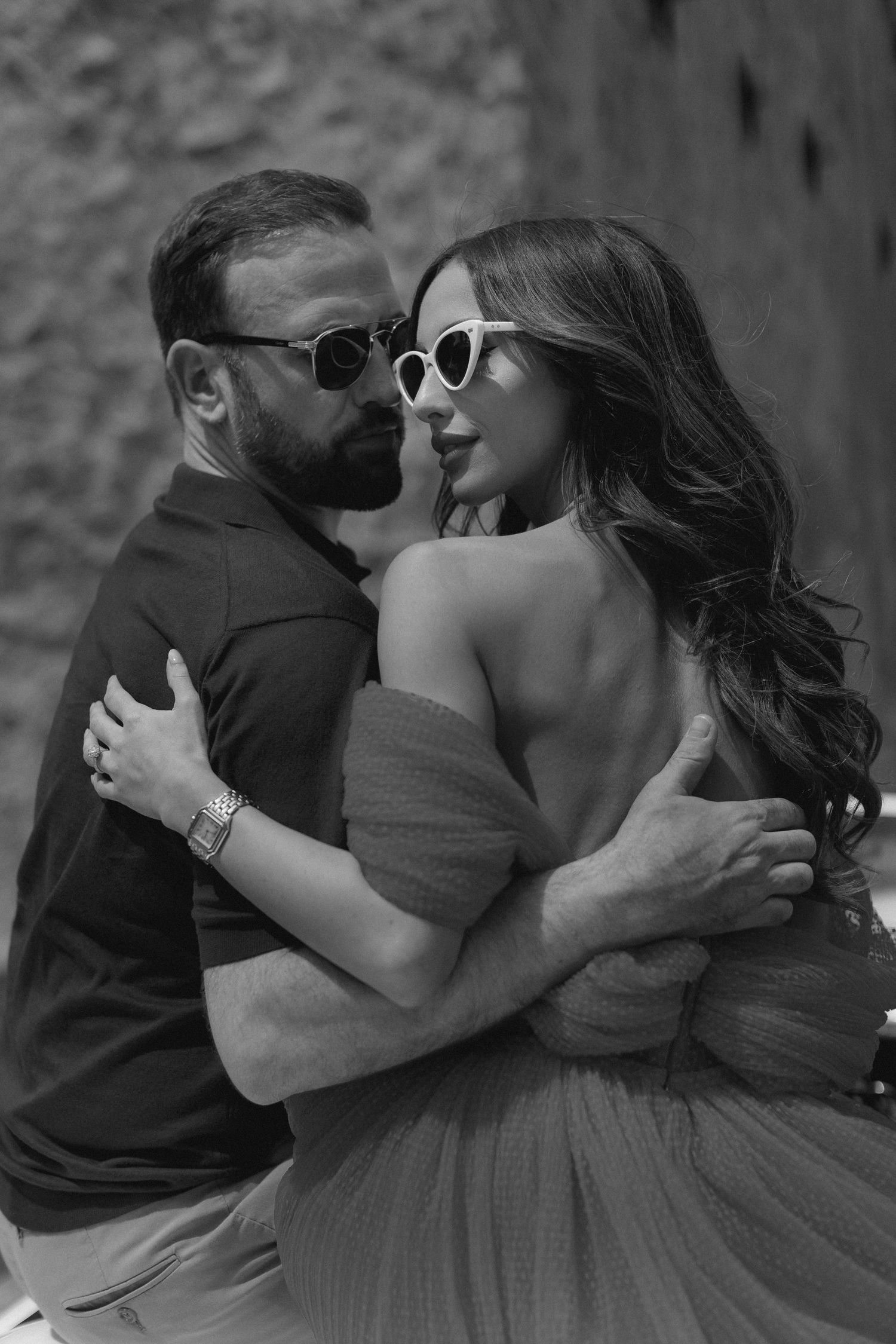 Romantic black and white portrait of a stylish couple in sunglasses embracing, with woman in off-shoulder dress.