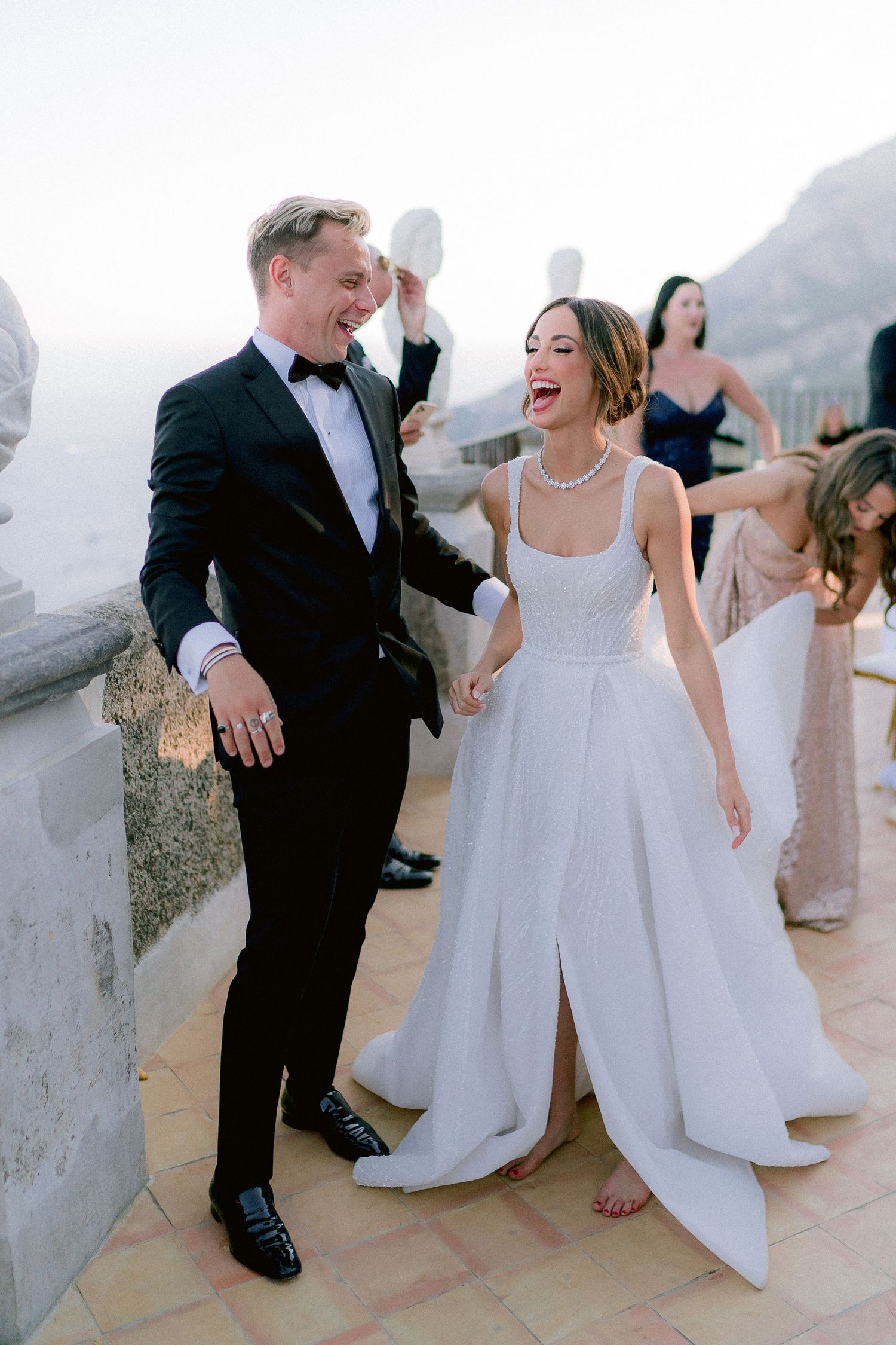 A joyful couple laughs together on a terrace overlooking scenic mountains, the bride in white and groom in classic tuxedo.