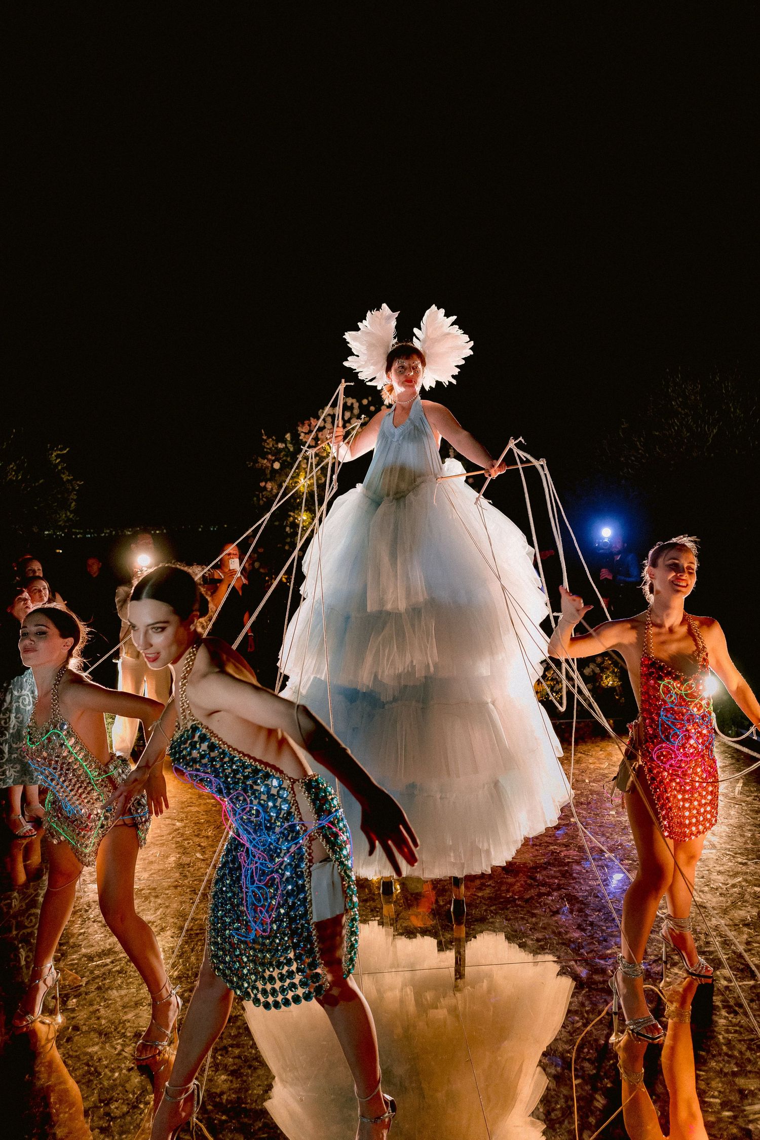 Performers in vibrant costumes dance and pose at night on a beach with figure in white ruffled dress elevated above others.