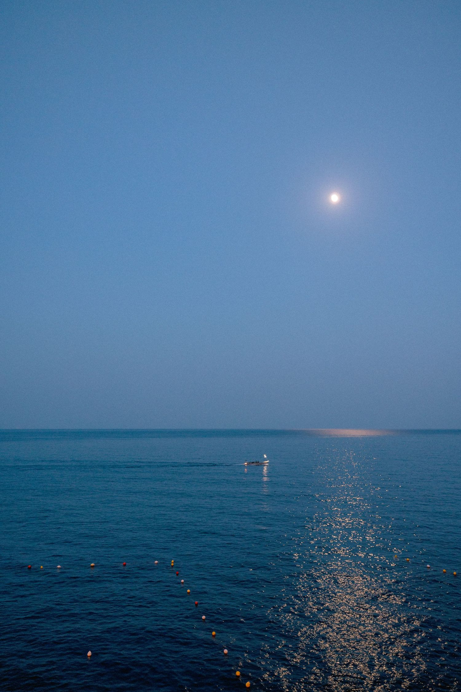 Moonlight reflects on calm blue ocean waters creating a glowing path across the surface while small lights float near horizon.