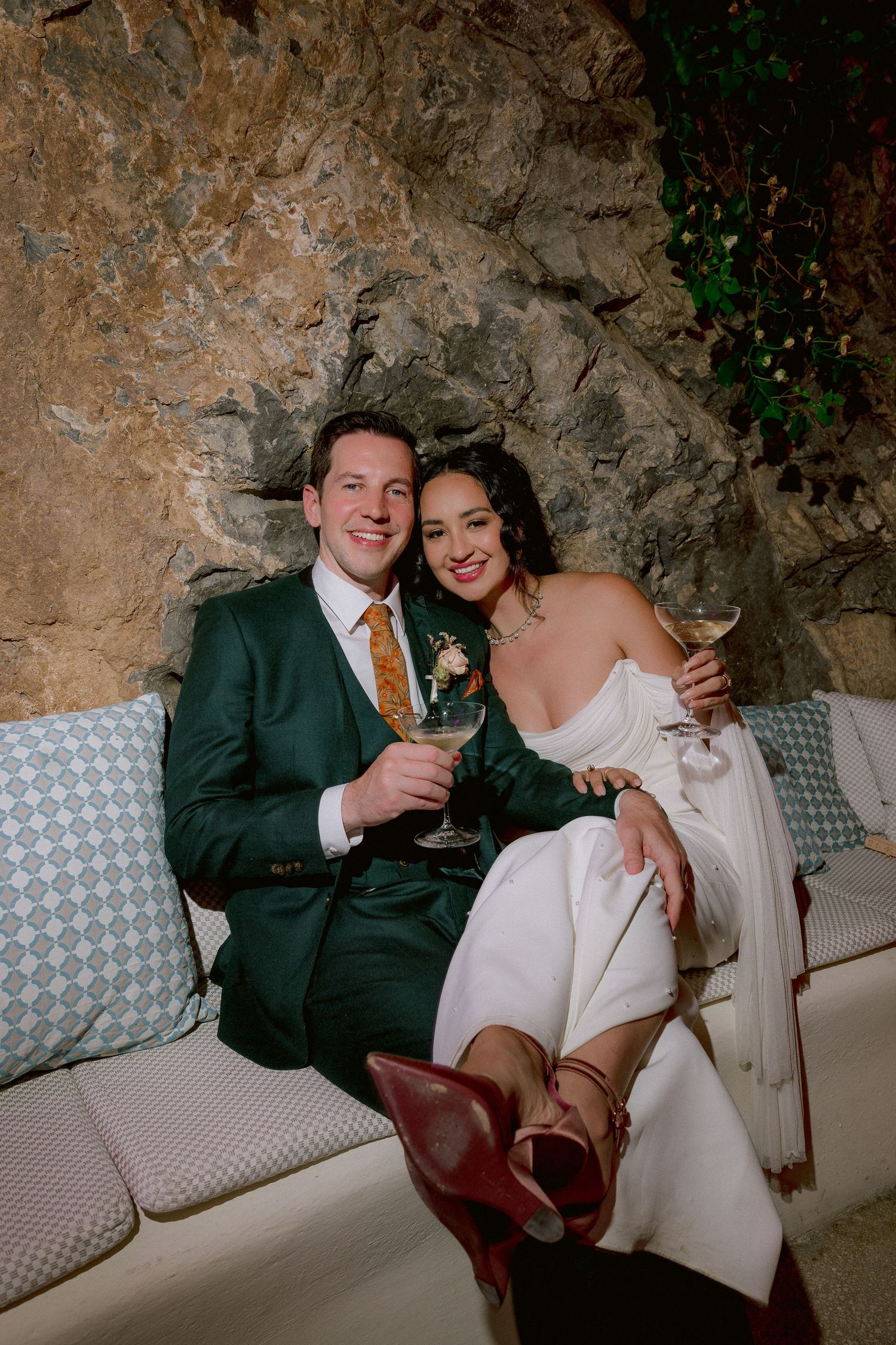 A couple in wedding attire sits together on a cushioned bench against a stone wall, holding champagne glasses.