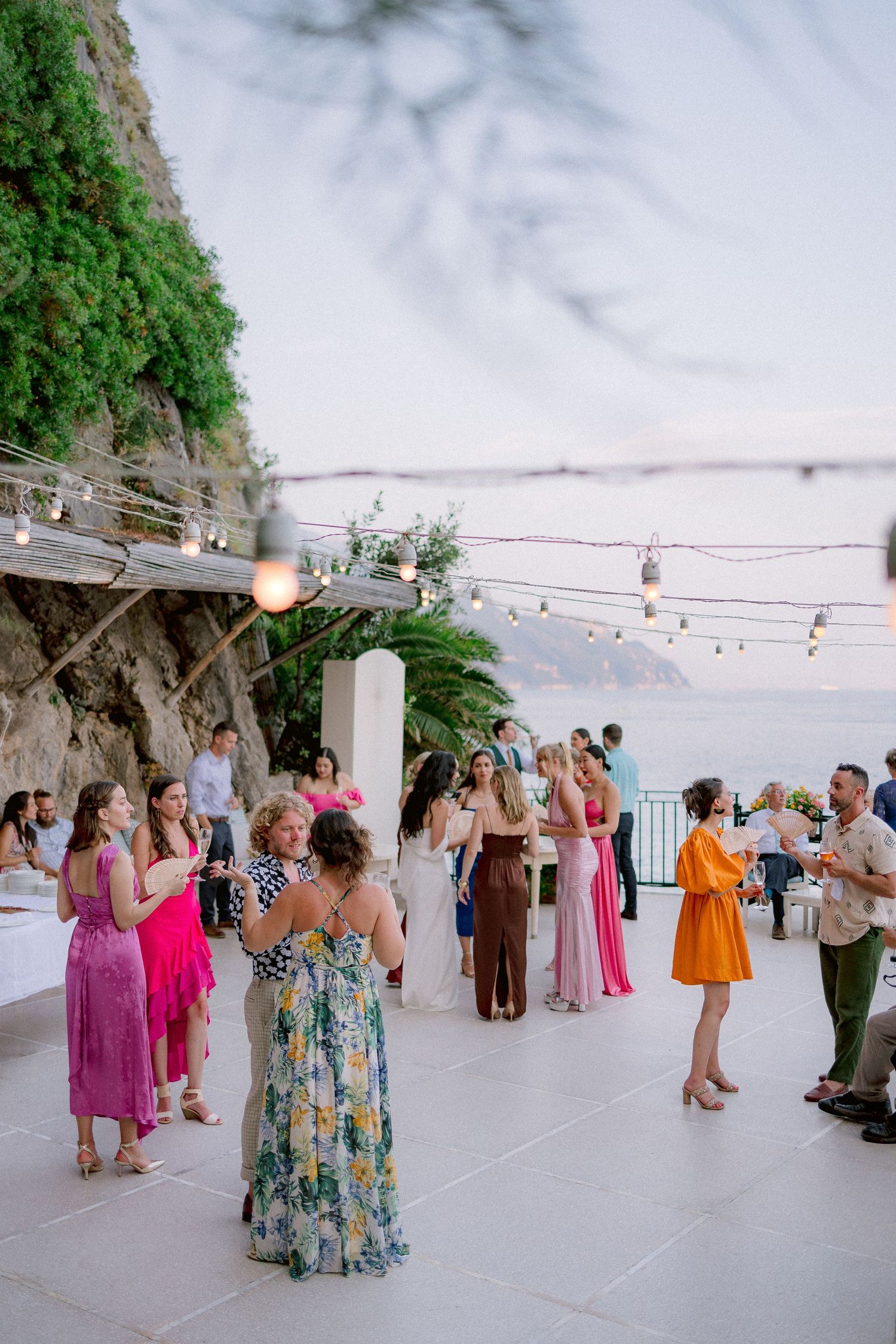 Party guests mingle and dance on an outdoor terrace with string lights overlooking a scenic coastal view at dusk.