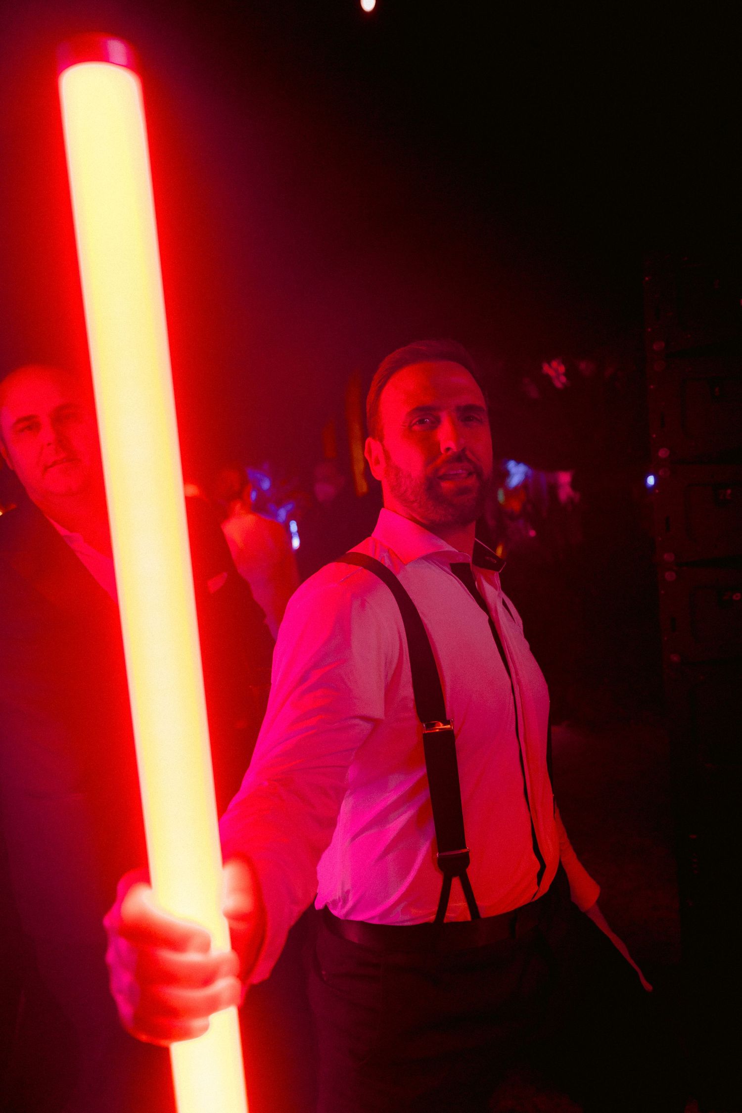 A performer in suspenders and white shirt holds up a glowing red light saber in a dark nightclub setting.