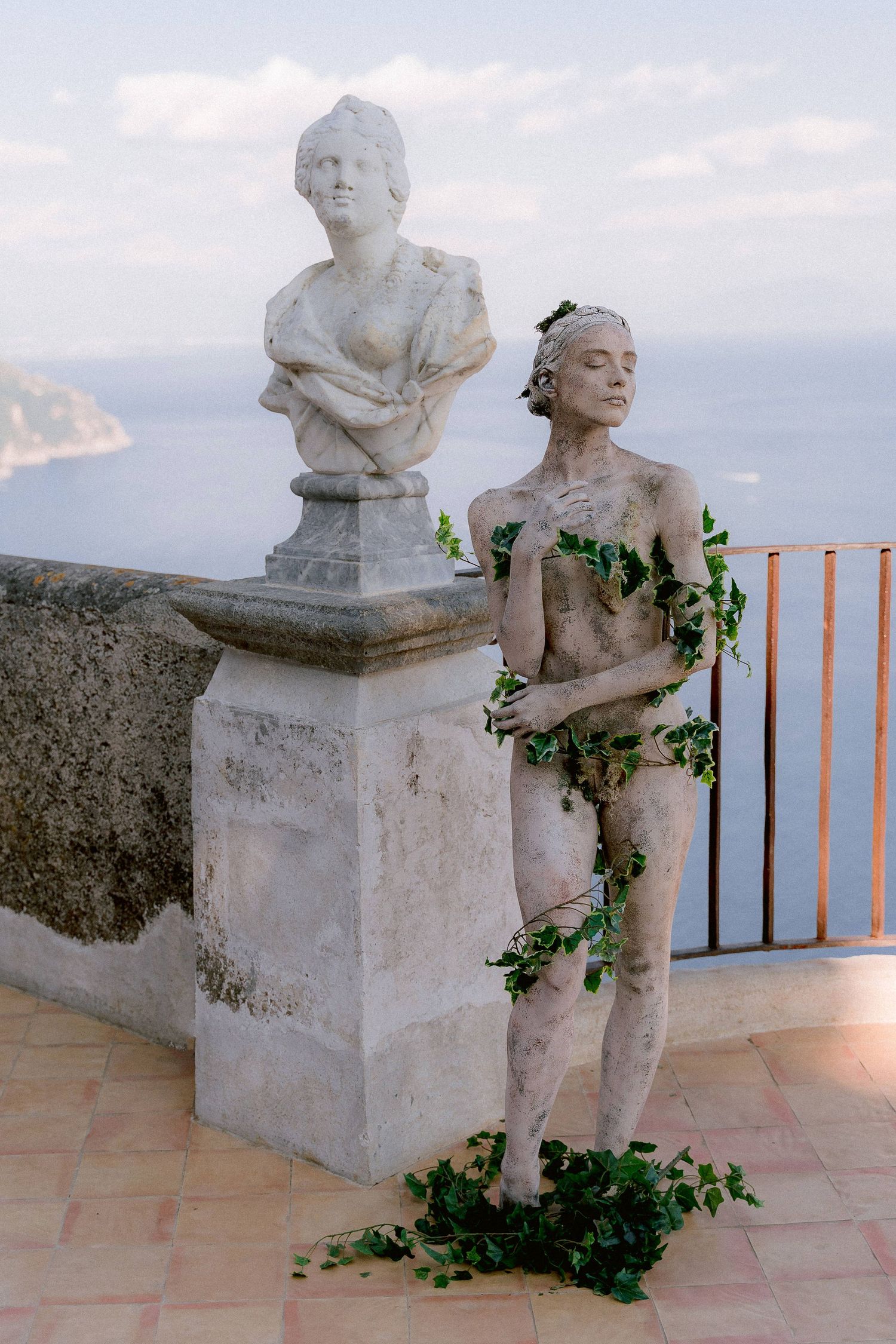 Two classical style statues on a Mediterranean balcony with climbing ivy vines and coastal views in the background.