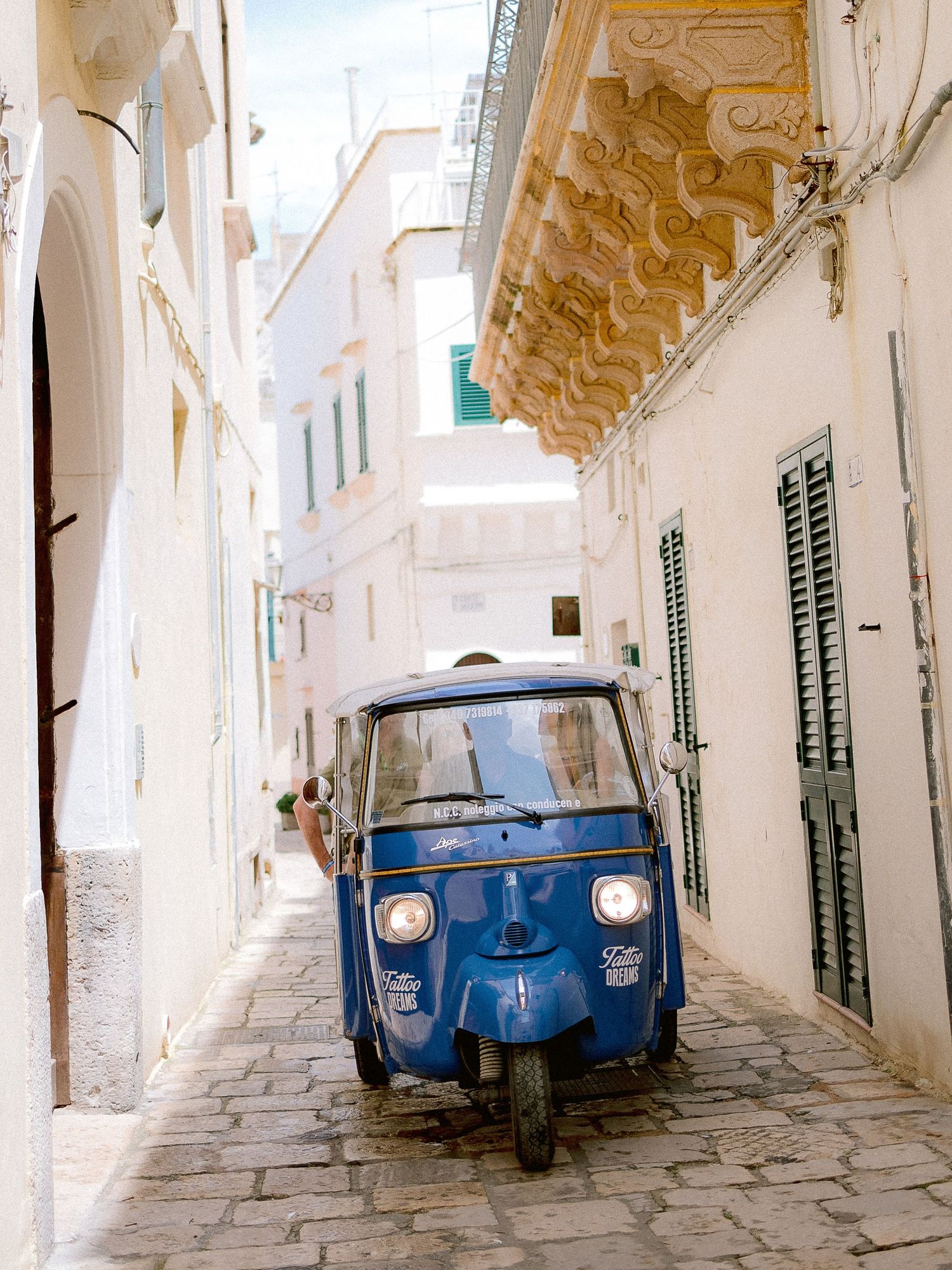 A vintage blue Piaggio Ape scooter parked in a narrow whitewashed alley with ornate architecture in Puglia.